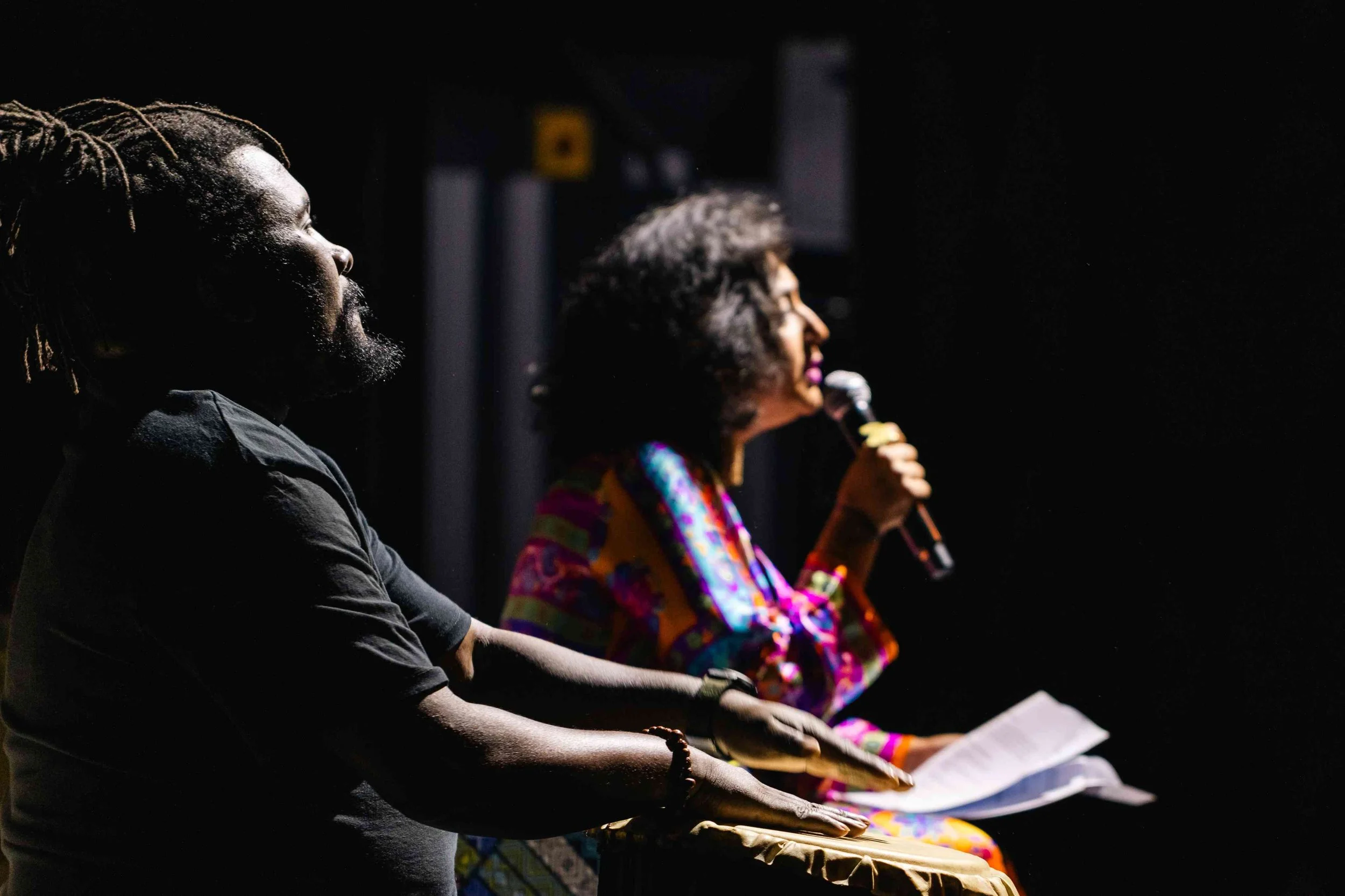 Two people are sitting in a dark room with a black background. The person on the left, with dreadlocks and beard, is facing to the right and resting their hands on a drum. The person on the right, with tied back hair, is holding a microphone and read