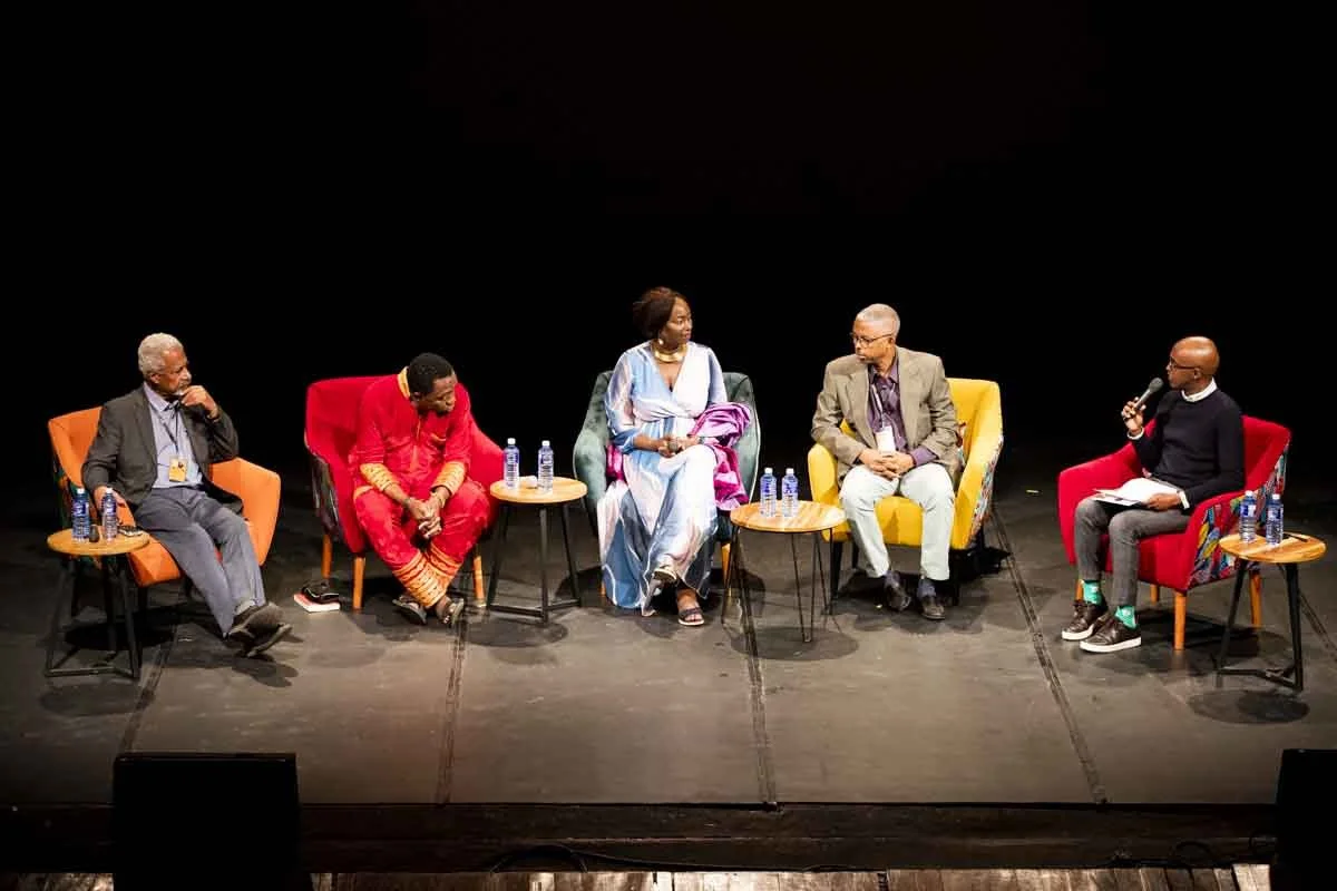 Six individuals seated on a stage engaged in a panel discussion, with black background and small tables with water bottles in front of them.