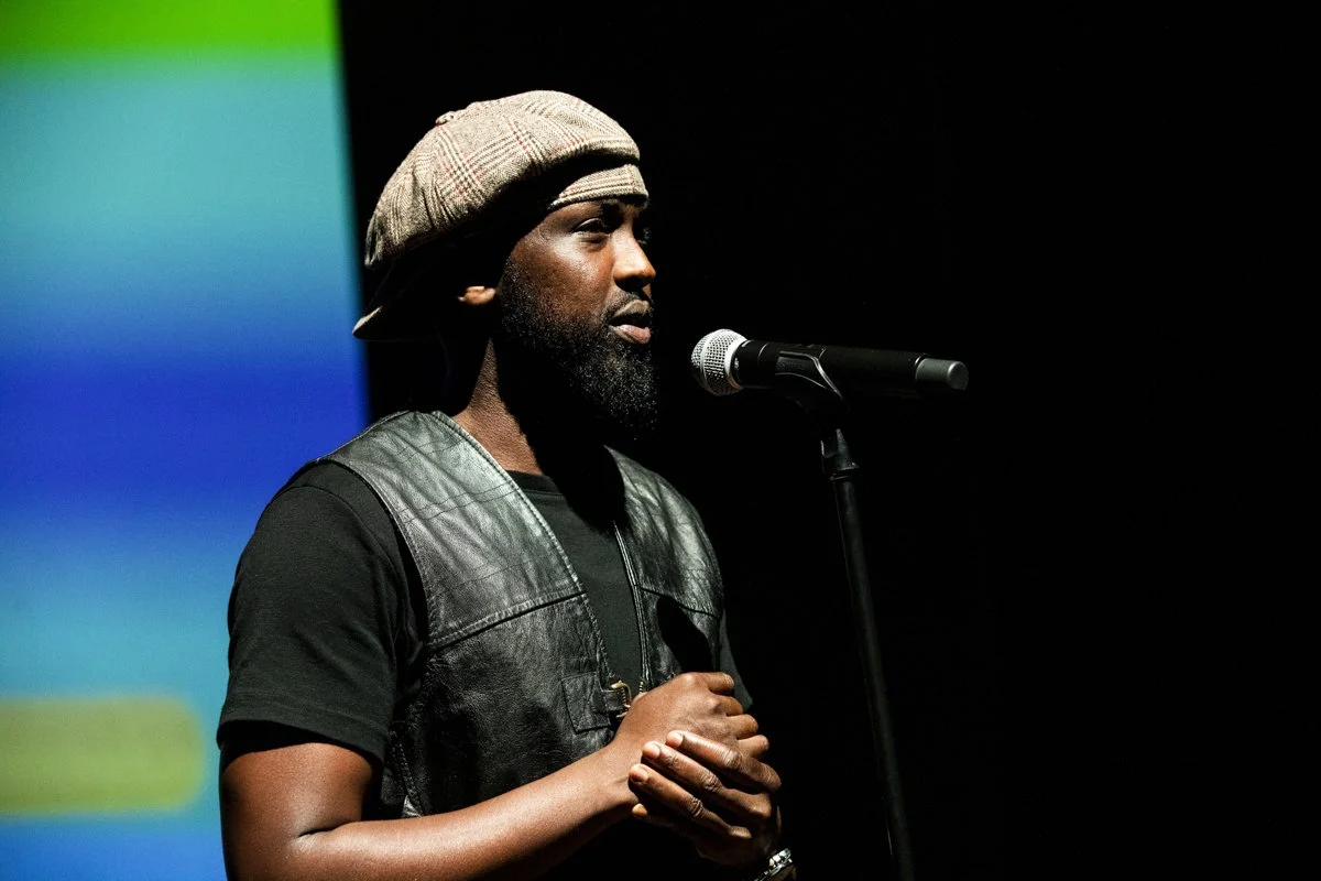Man with a beard and a cap standing in front of a microphone on a dark background, with a colorful gradient backdrop on the left side.