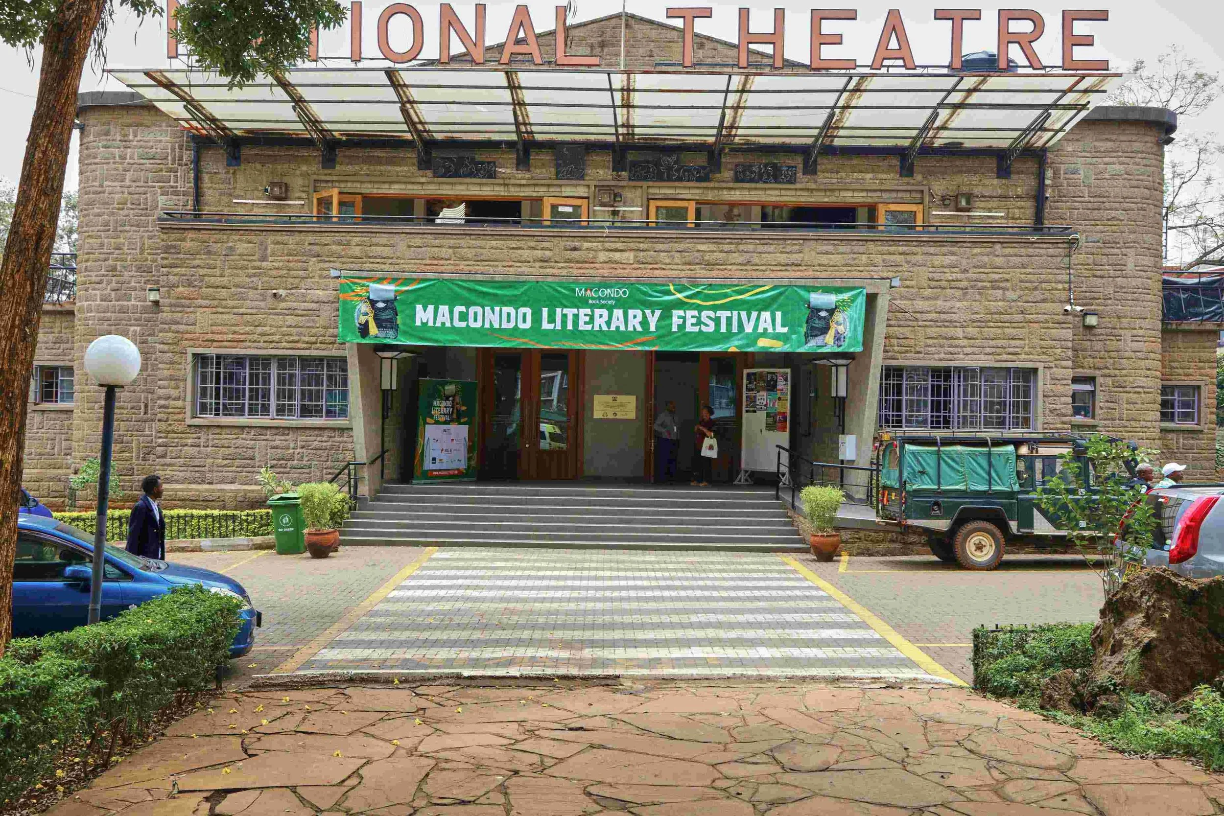 The entrance of a stone building with a sign for the Macondo Literary Festival, featuring a green banner, stairs leading up to the door, and parked vehicles in front.