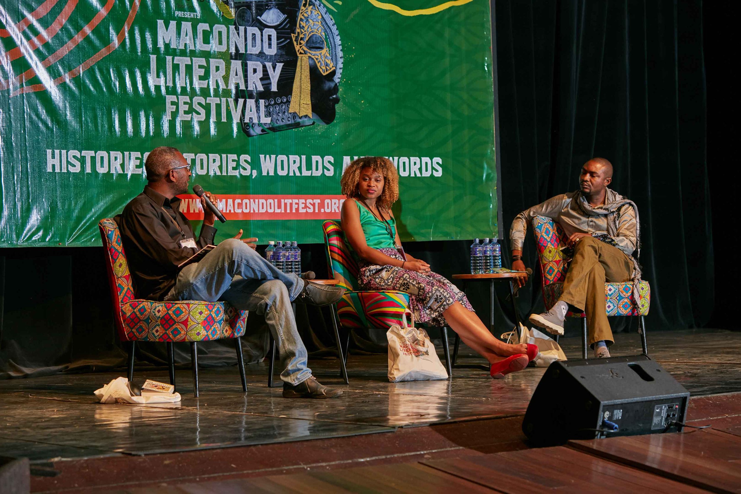 Three people seated on stage at the Macondo Literary Festival, with a green backdrop featuring the festival's logo and slogan. The man on the left holds a microphone and is speaking, the woman in the center looks on, and the man on the right listens 