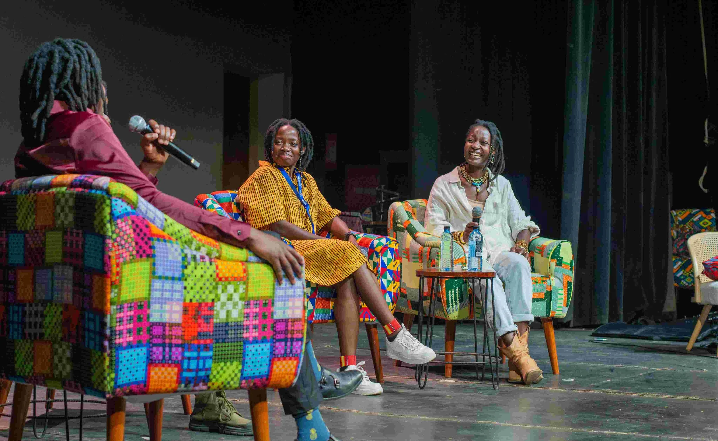 Three women seated on colorful patchwork chairs on a stage, each holding a microphone, engaged in conversation during a panel discussion.