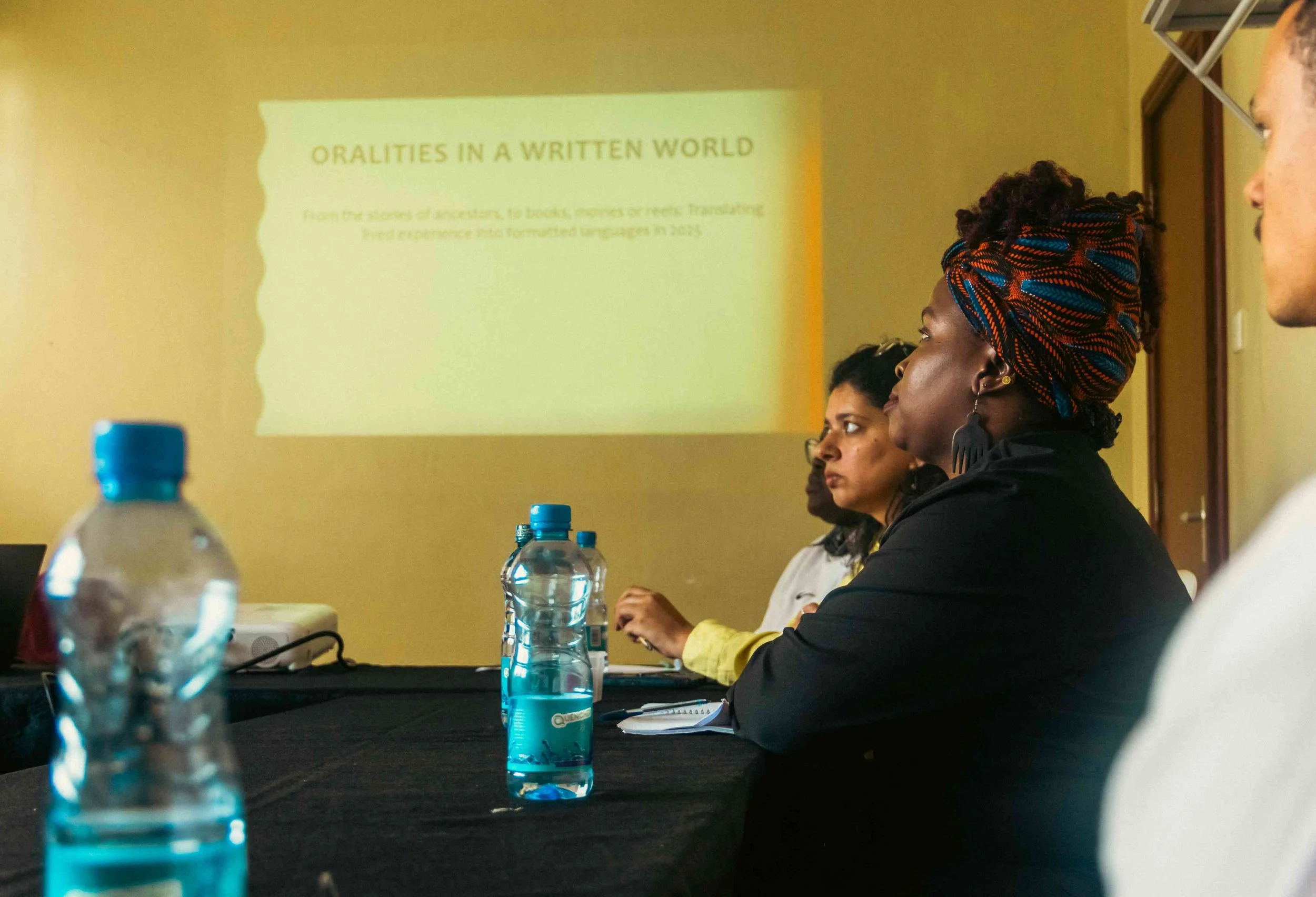 Women attending a presentation about orality in a written world, with bottles of water on the table and a projected slide on the wall.