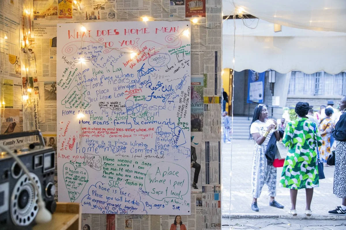A whiteboard with colorful handwritten messages and drawings about the meaning of home, surrounded by newspaper pages, with a string of lights, inside a tent or booth. Outside, people are gathered and talking.