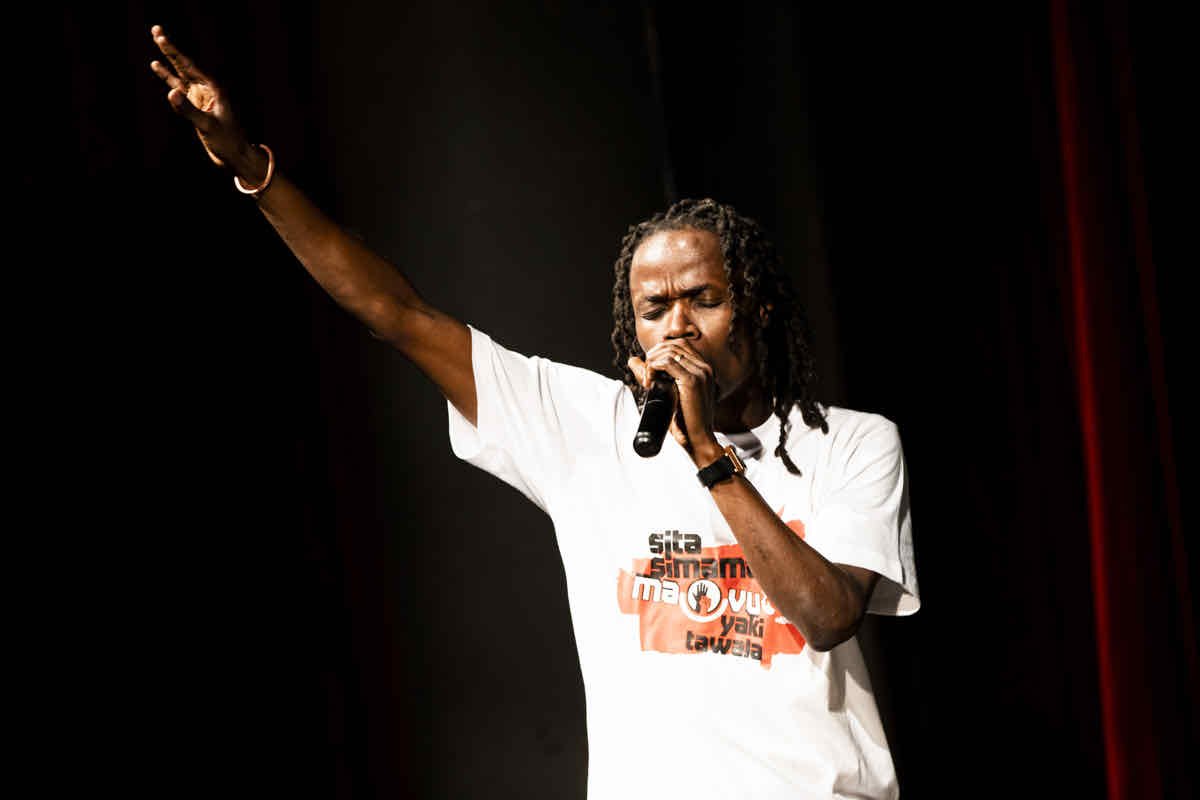 A man with dreadlocks singing into a microphone with eyes closed, raising one arm in the air, on stage with a dark background.