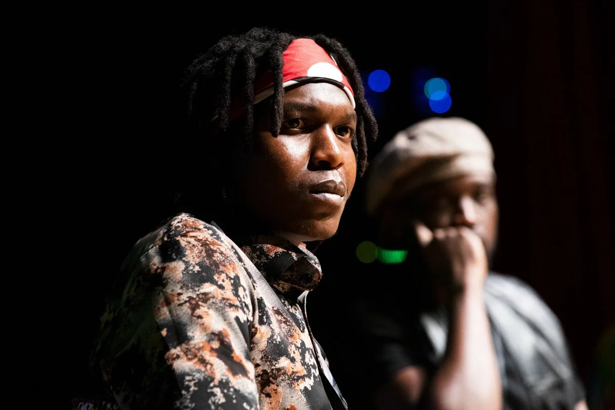 A young man with dreadlocks wearing a red headband and a patterned shirt looks directly at the camera, with a man in the background wearing a headwrap and holding his hand over his mouth, in low lighting with blurred colorful lights.