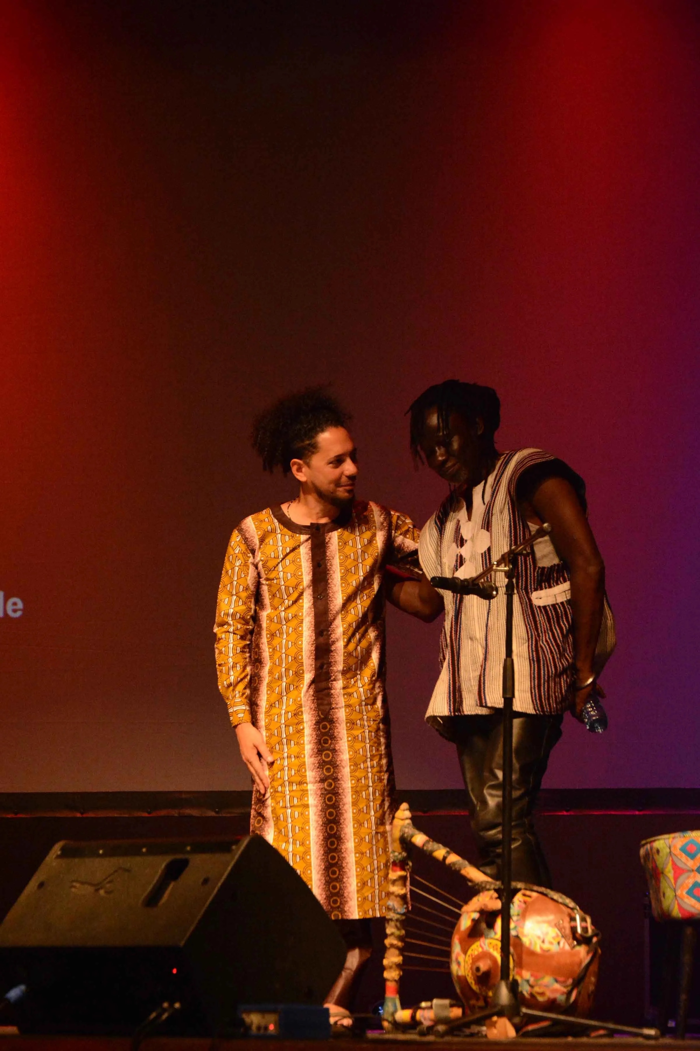 Two men on stage, one wearing a colorful patterned dress, and the other wearing a striped shirt, are interacting with each other. They are standing near a microphone with a colorful basket or drum on the floor beside them, and a speaker is visible in