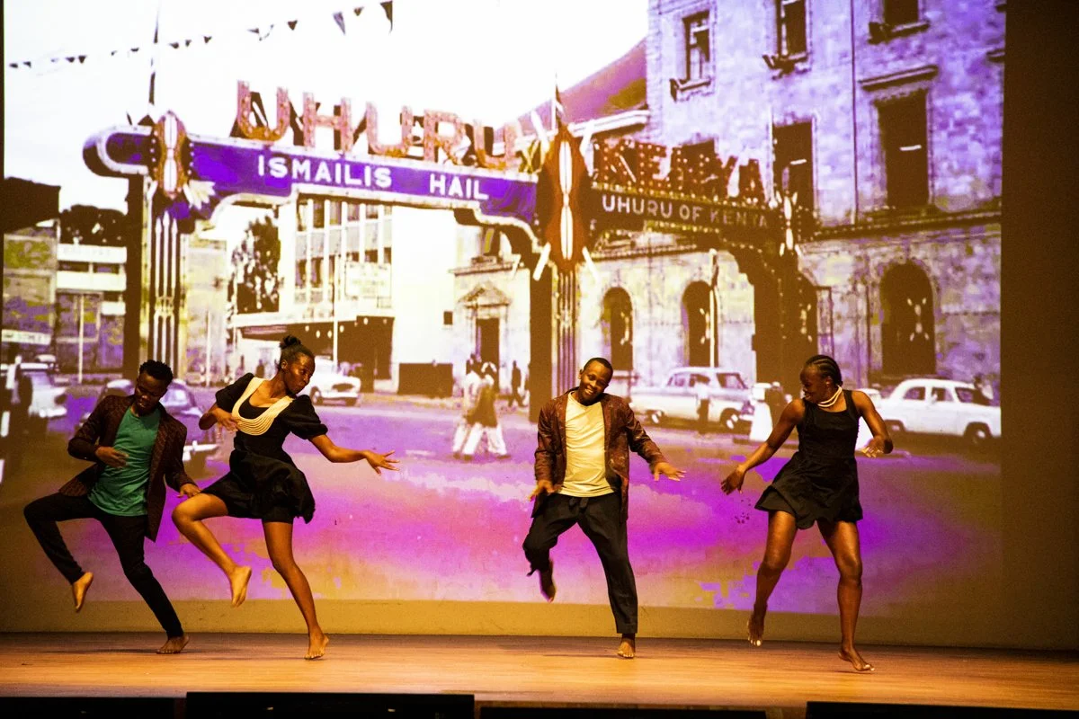 Four dancers performing on stage with a vintage cityscape backdrop featuring a sign that reads 'FURRY,' 'IS MAILS HAIL,' and 'UNION OF KENYA.'