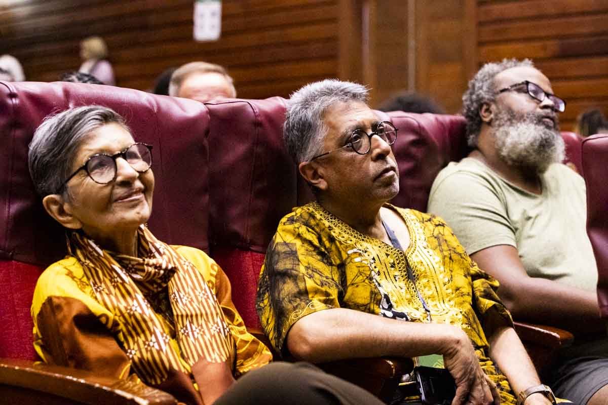 Three elderly individuals seated in auditorium chairs, listening attentively, with two women and one man, all wearing glasses and colorful clothing, in a wood-paneled room.