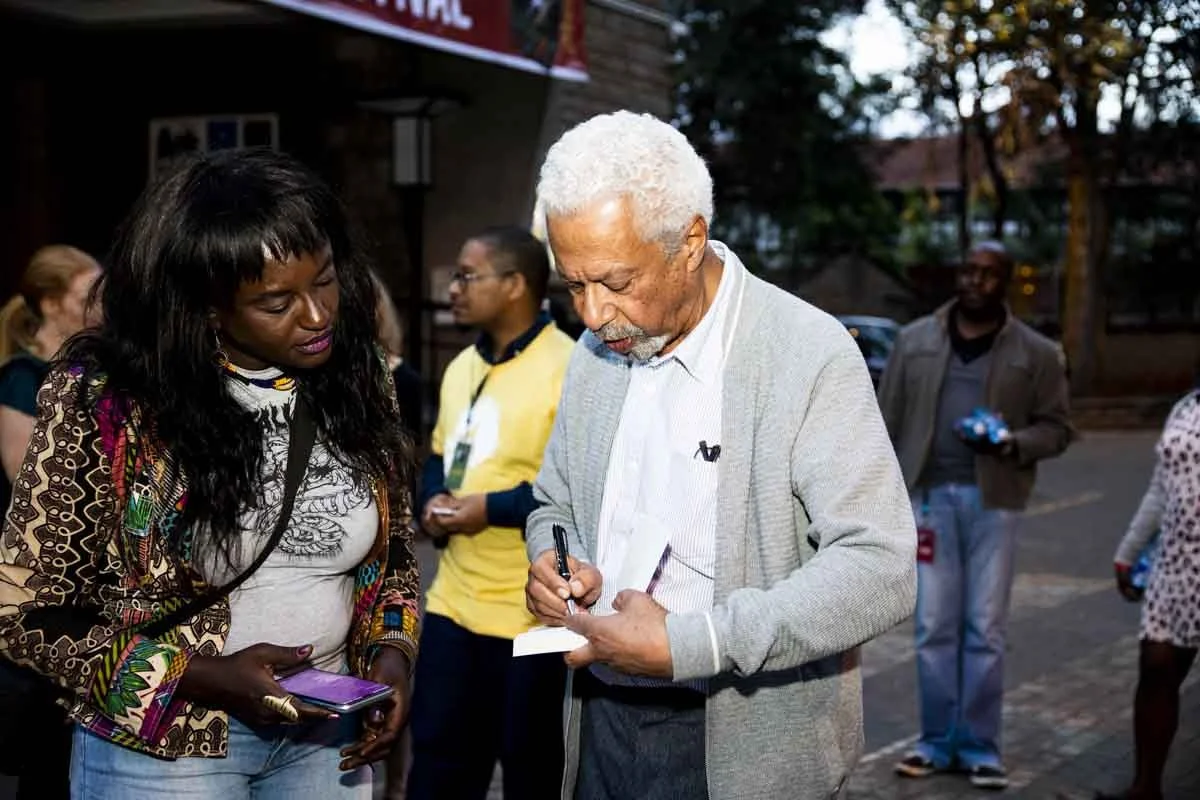 An elderly man with white hair is signing a book for a woman with long dark hair holding a phone. Several people stand around in the background outside on a paved area, with trees and buildings visible.