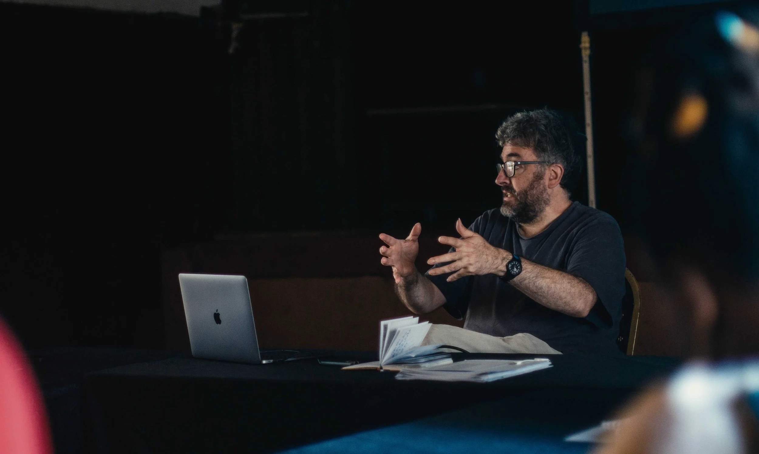 Man with glasses and beard gesturing while sitting at table with open laptop and papers in a dark room