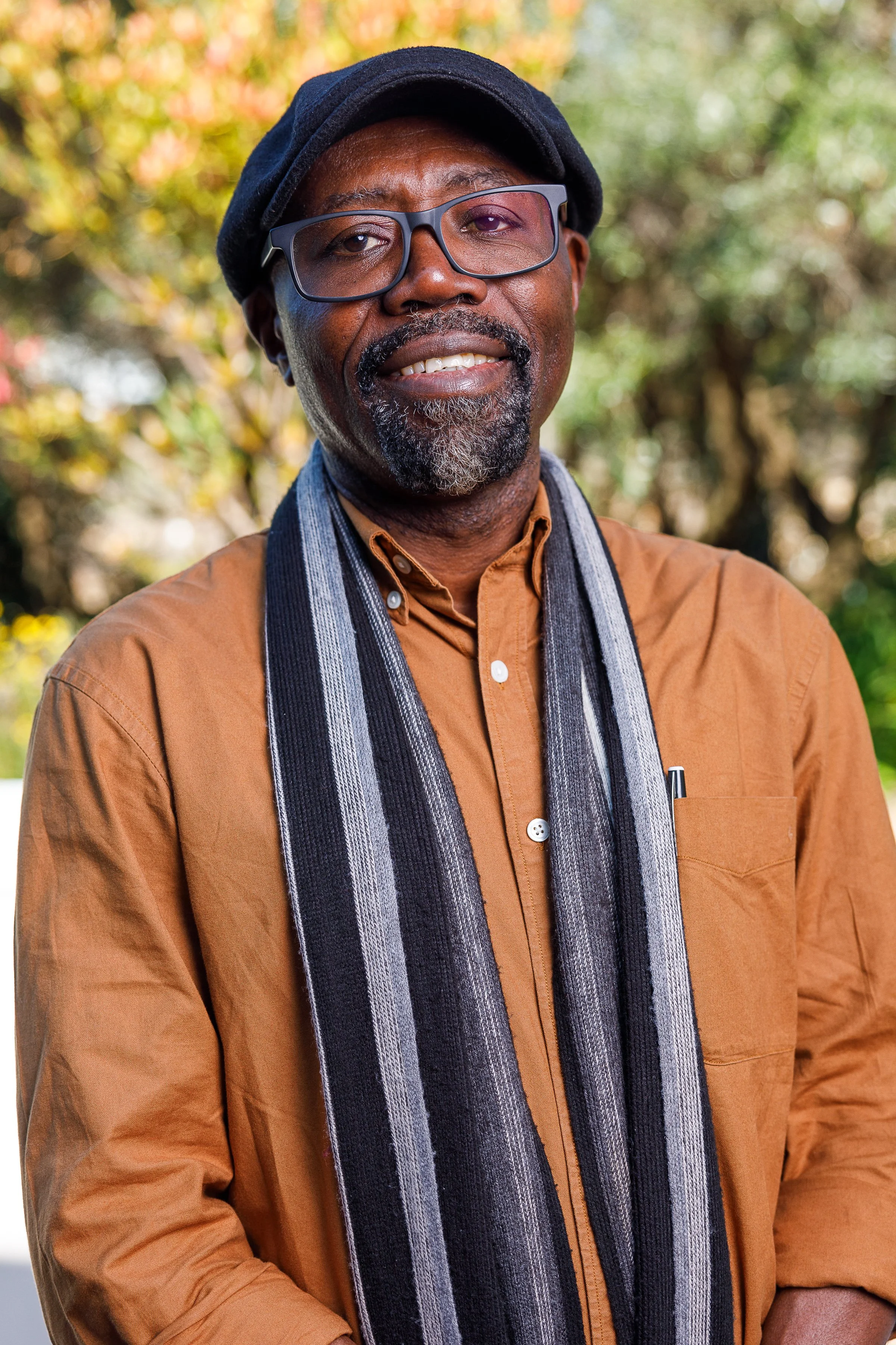 A middle-aged man with glasses and a beard, wearing a black cap, a brown shirt, and a striped scarf, standing outdoors with autumn foliage in the background.