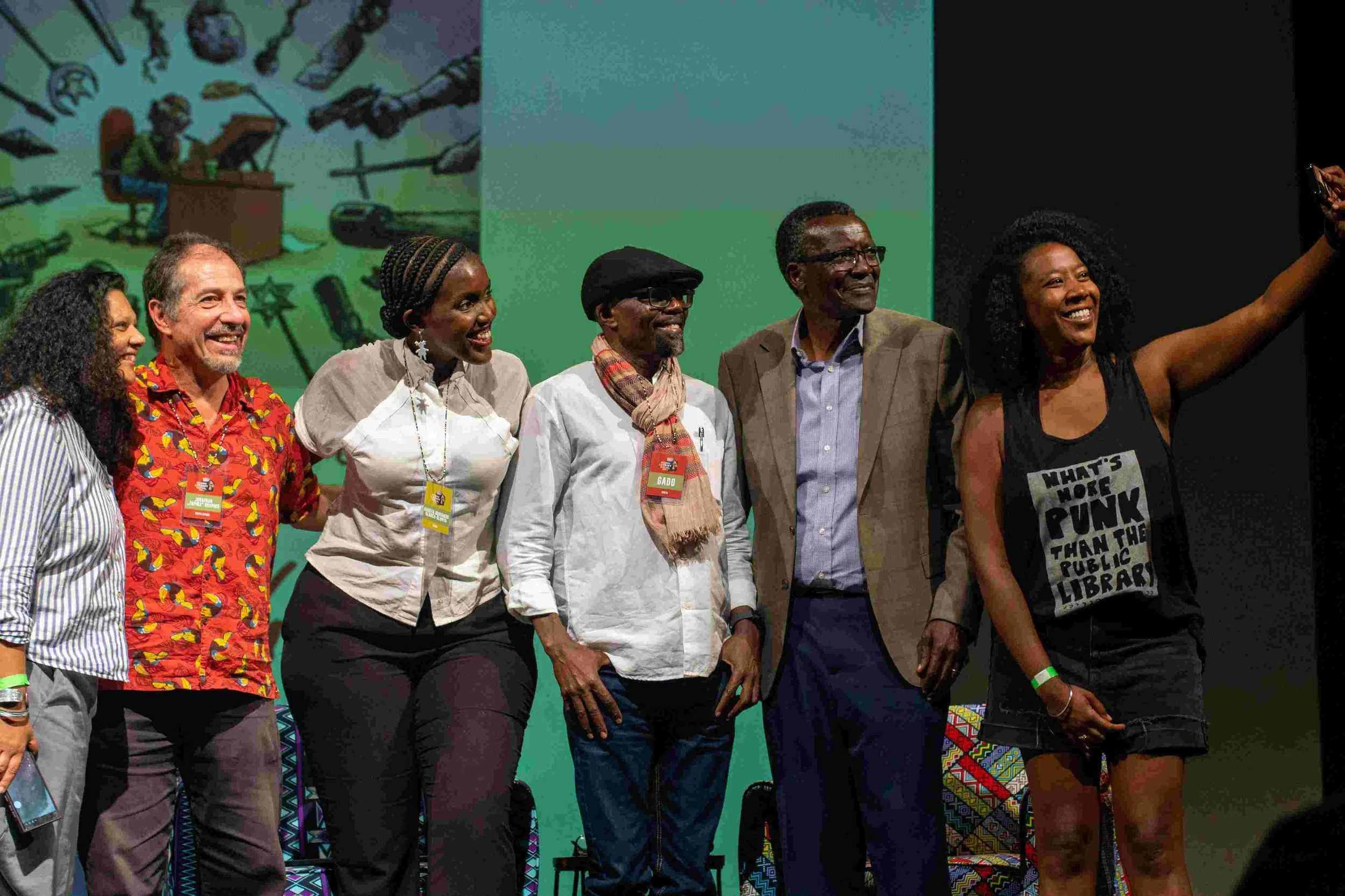 Six diverse individuals standing on stage, smiling, and taking a selfie. Behind them, a colorful mural features a person playing a piano surrounded by various musical instruments.