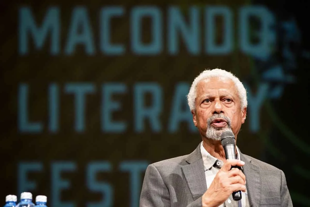 An elderly man with white hair and beard speaking into a microphone on stage, with a background reading 'Macondo Literary Festival'.