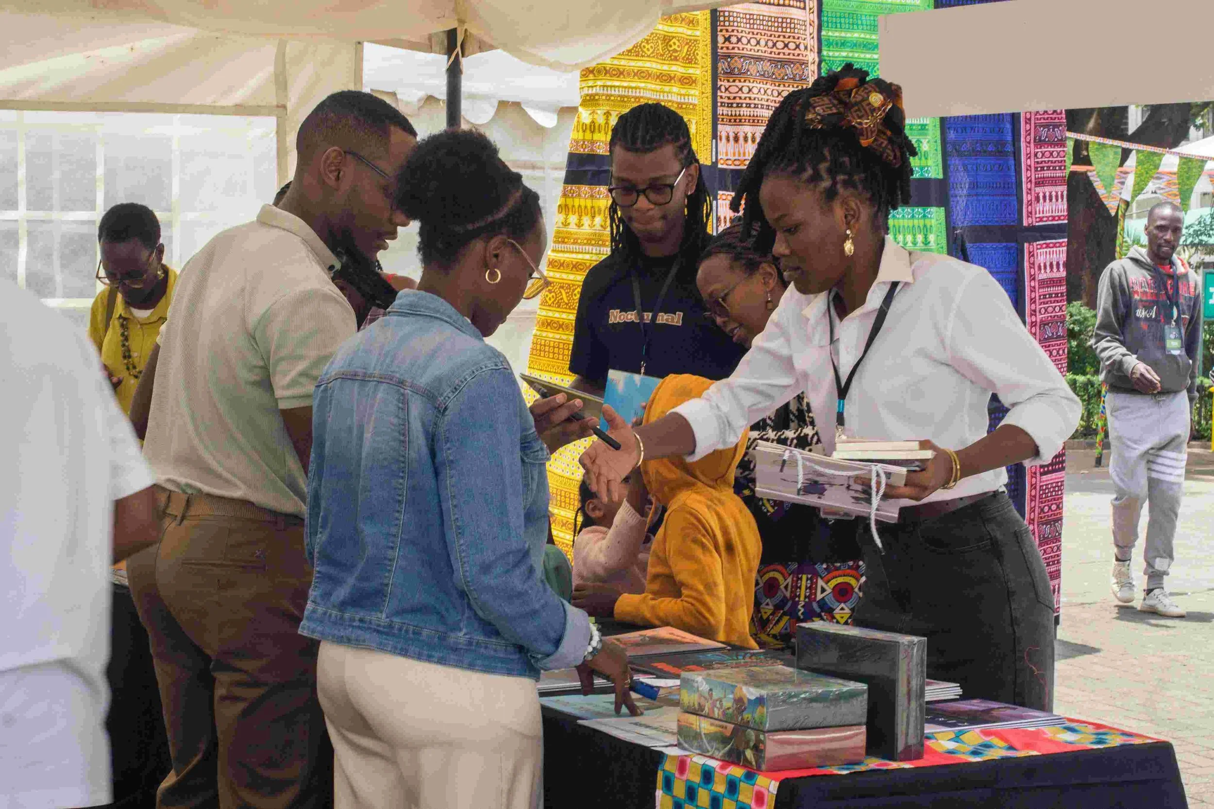 Group of people gathered around a table at an outdoor cultural event, with colorful traditional fabric decorations in the background. They are looking at books and engaging with each other.