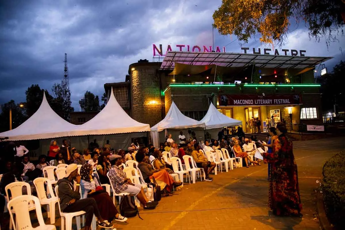 People attending the Macondo Literary Festival outside the National Theatre, seated on white chairs under white tents, with a building and neon signs in the background during evening.