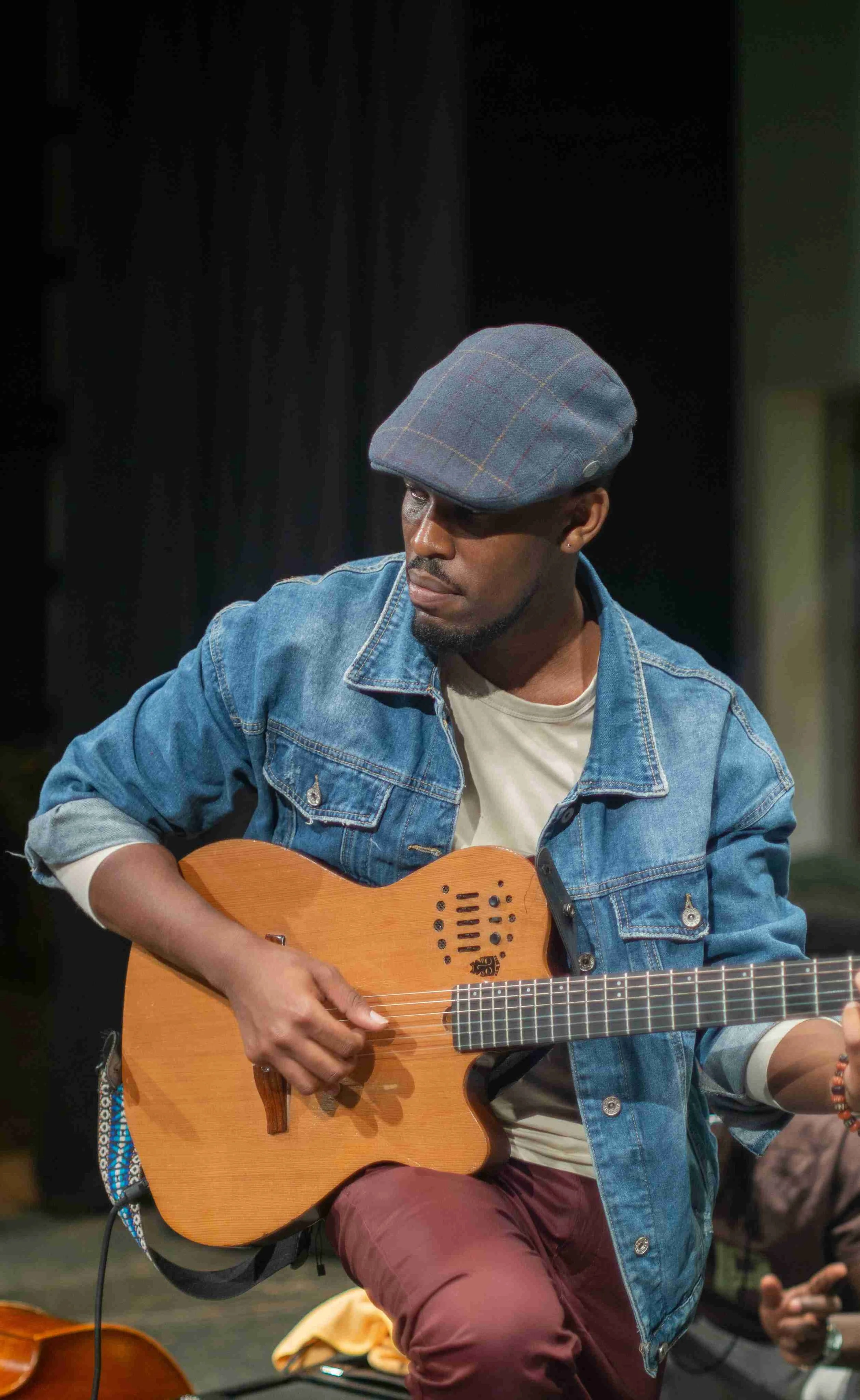 A man wearing a plaid cap, denim jacket, and beige shirt playing an acoustic guitar.