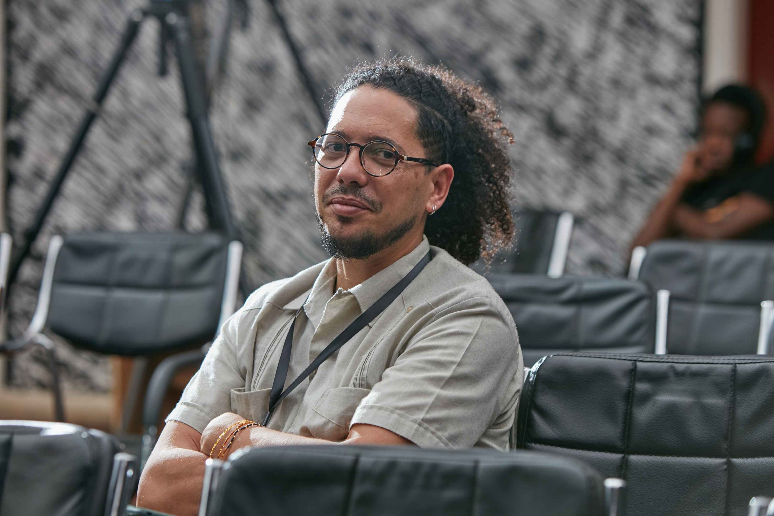 A man with glasses, earrings, and curly hair sits with arms crossed in a room with black chairs and a textured wall, with a woman blurred in the background.