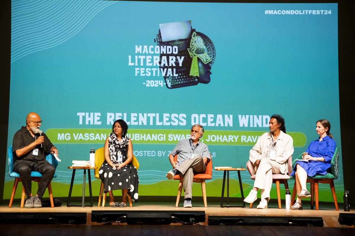 Panel discussion at the Macondo Literary Festival 2024 with five speakers seated on stage, speaking into microphones, with a large blue background screen displaying the festival's event title and topic.