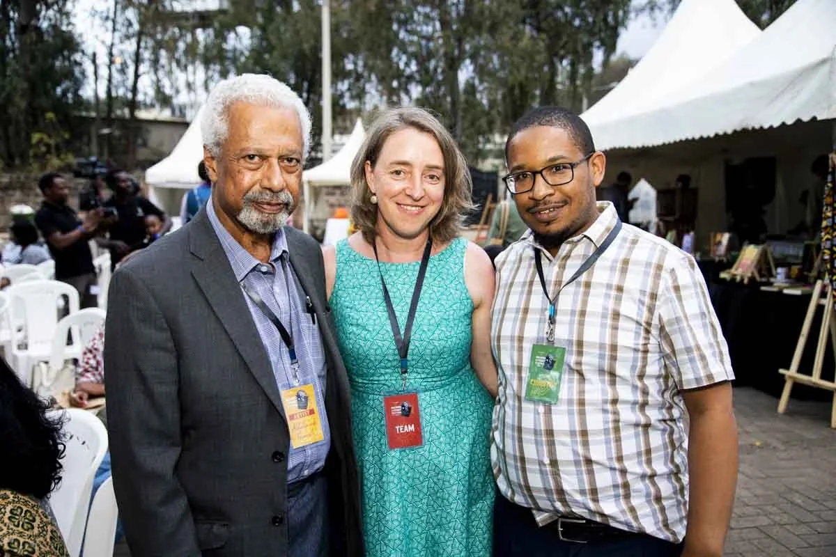 Three individuals at an outdoor event, standing closely together and smiling at the camera. They are wearing badges, with tents and trees in the background.