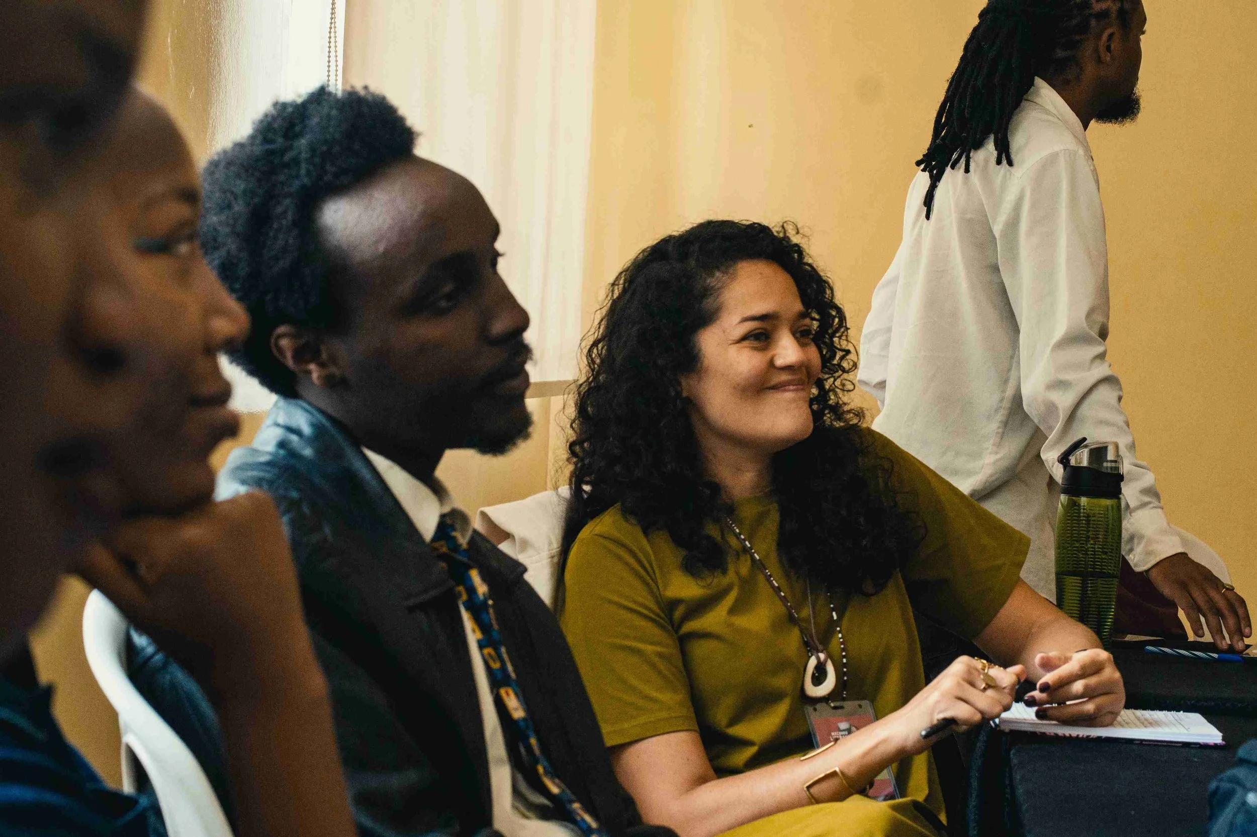 A group of diverse people sitting and listening in a meeting or conference, with one woman smiling and holding a pen.