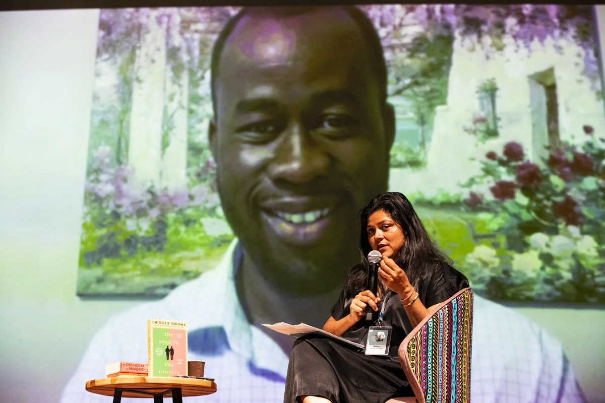 A woman with dark hair and a black dress sitting on a colorful chair, holding a microphone while speaking. Beside her on a small round table are several books. Behind her, a large screen displays a smiling man with short hair, wearing a white shirt, 