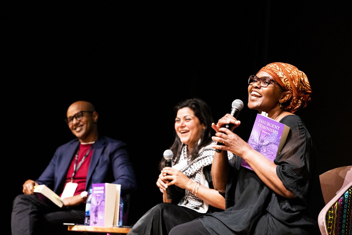 Three diverse women sitting on stage, speaking into microphones, smiling, and holding books titled "Insurgent Feminisms" during a panel discussion.