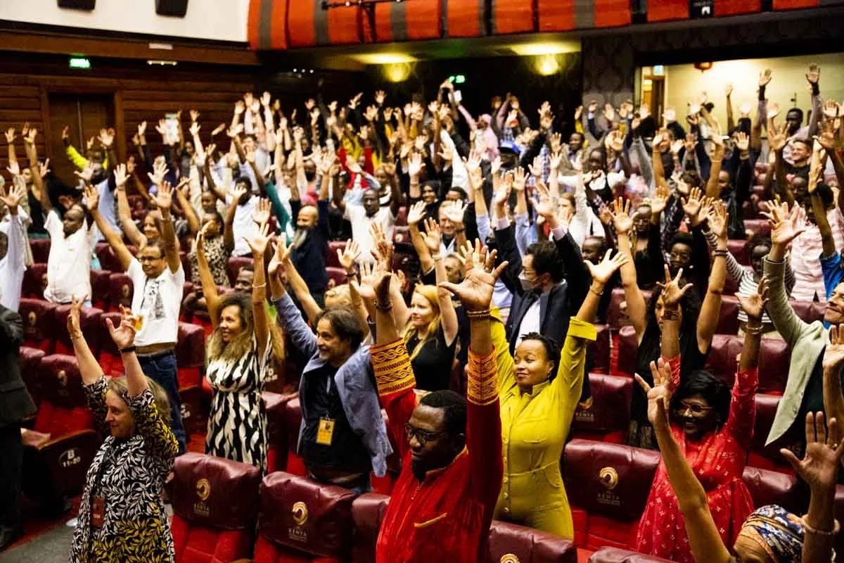 A large group of people attending a conference or event, raising their hands in a cheer or gesture of celebration in an auditorium setting.