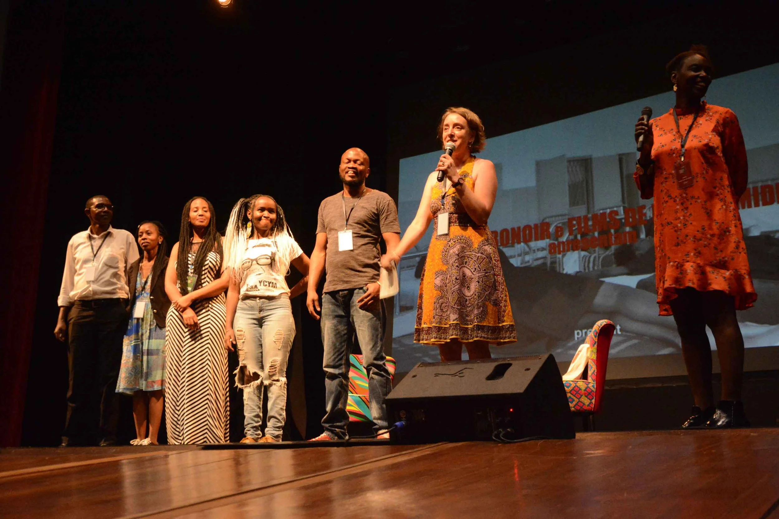 Group of seven people standing on stage, some holding microphones, with a large screen behind them displaying text and an image of a building.