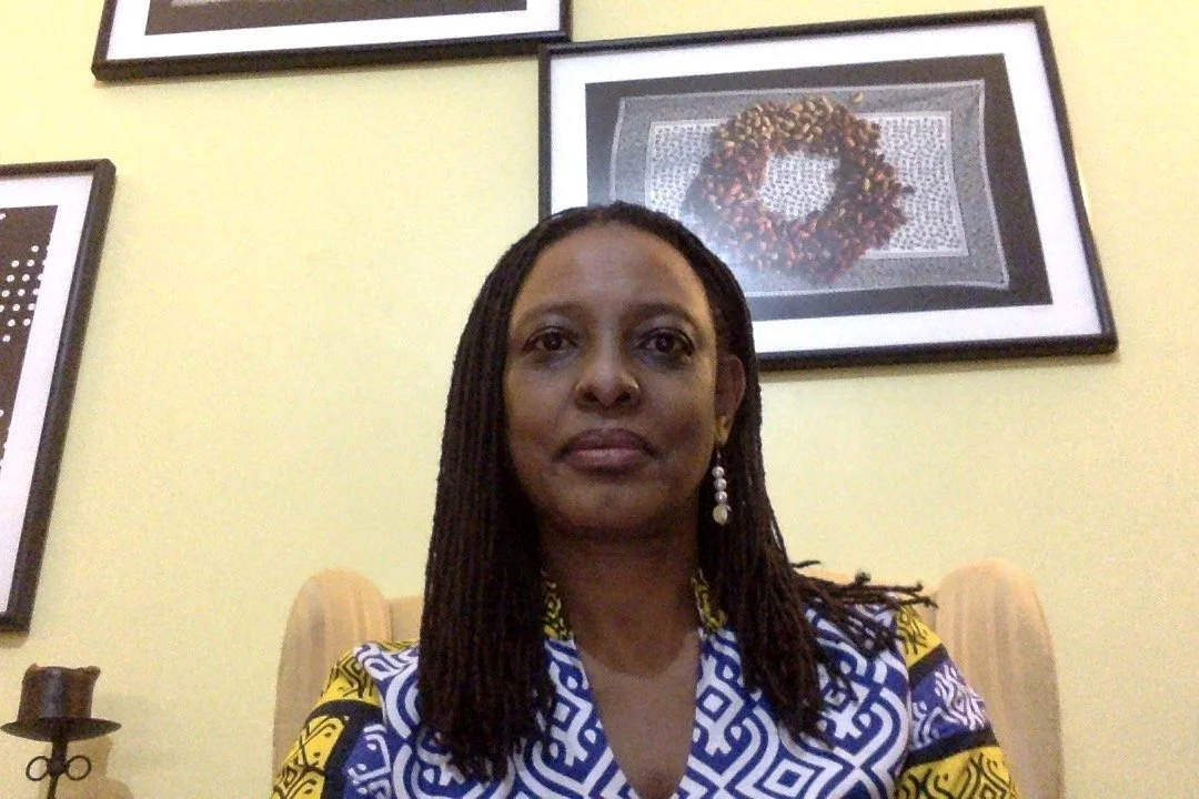A woman with dark braided hair, wearing earrings and a patterned dress, sitting on a beige chair in front of a yellow wall with framed artwork, including a wreath, hanging behind her.