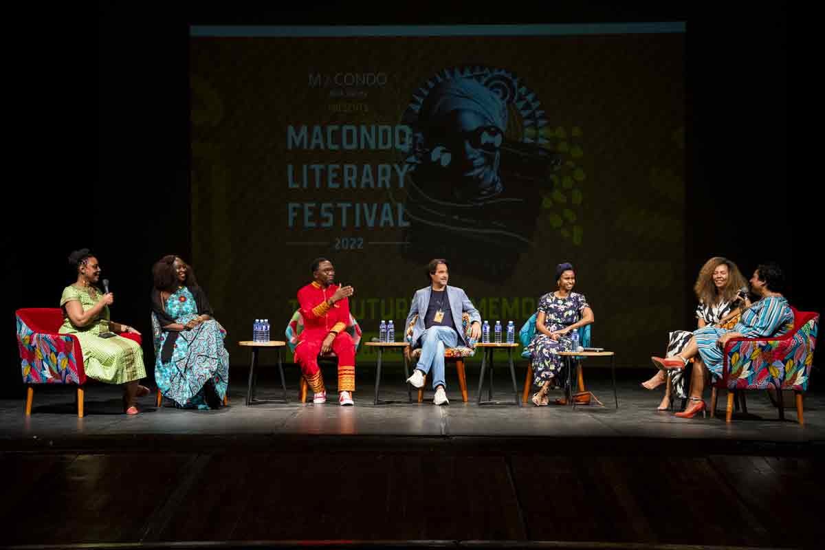 Panel of seven diverse women and one man sitting on stage in colorful chairs, participating in a discussion at the Macondo Literary Festival 2022, with a large screen in the background displaying the event logo.