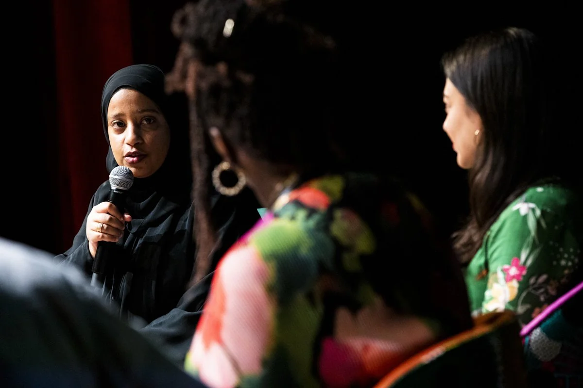 A woman in a hijab holding a microphone speaking at a panel discussion, with two women listening attentively.