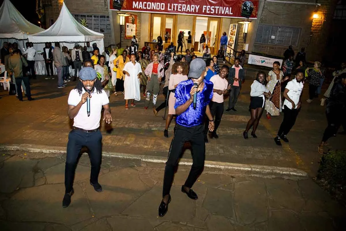People dancing and socializing outside the Macondo Literary Festival at night, with two men singing or speaking into microphones in the foreground.