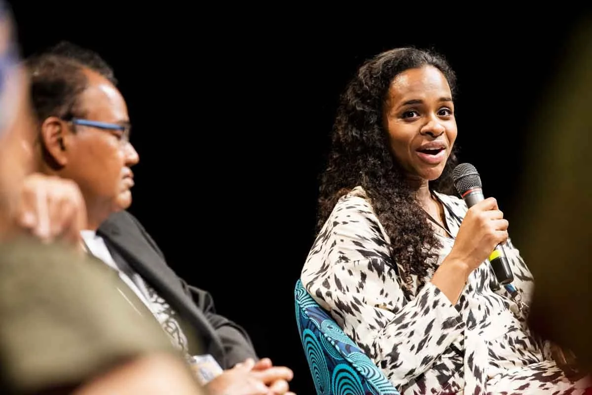 A woman with long curly hair holding a microphone and speaking at a panel or discussion event, with a man in glasses and a suit listening or waiting to speak next to her.