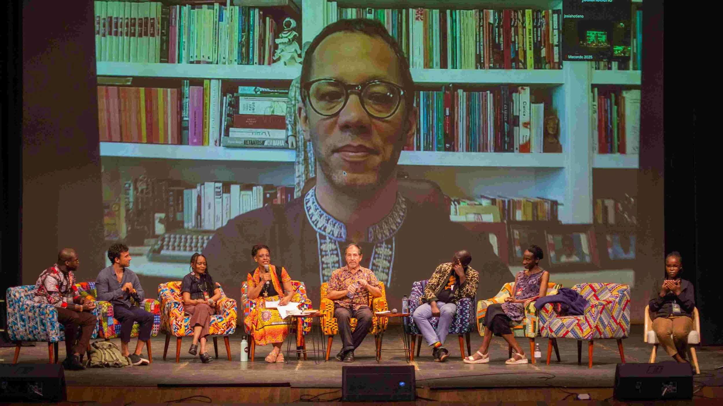 Panel discussion on stage with seven speakers seated in colorful chairs, large screen behind showing a person with glasses in front of a bookshelf. Panelists include men and women dressed in traditional and casual attire.