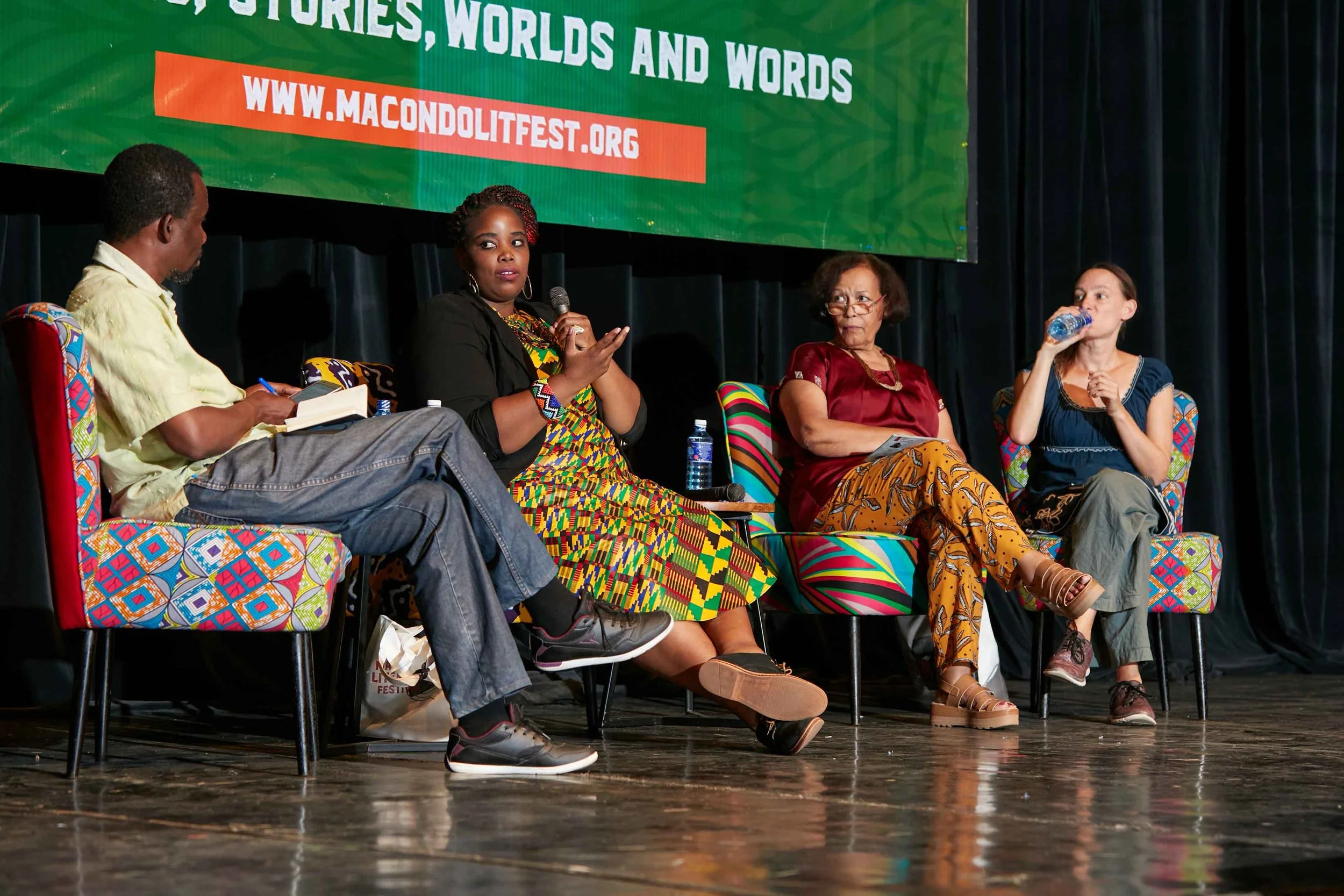 Four people seated on stage participating in a panel discussion at an event with a green banner overhead. The banner reads 'Stories, Worlds and Words' with a website address. The woman second from the left is speaking, holding a microphone, wearing a