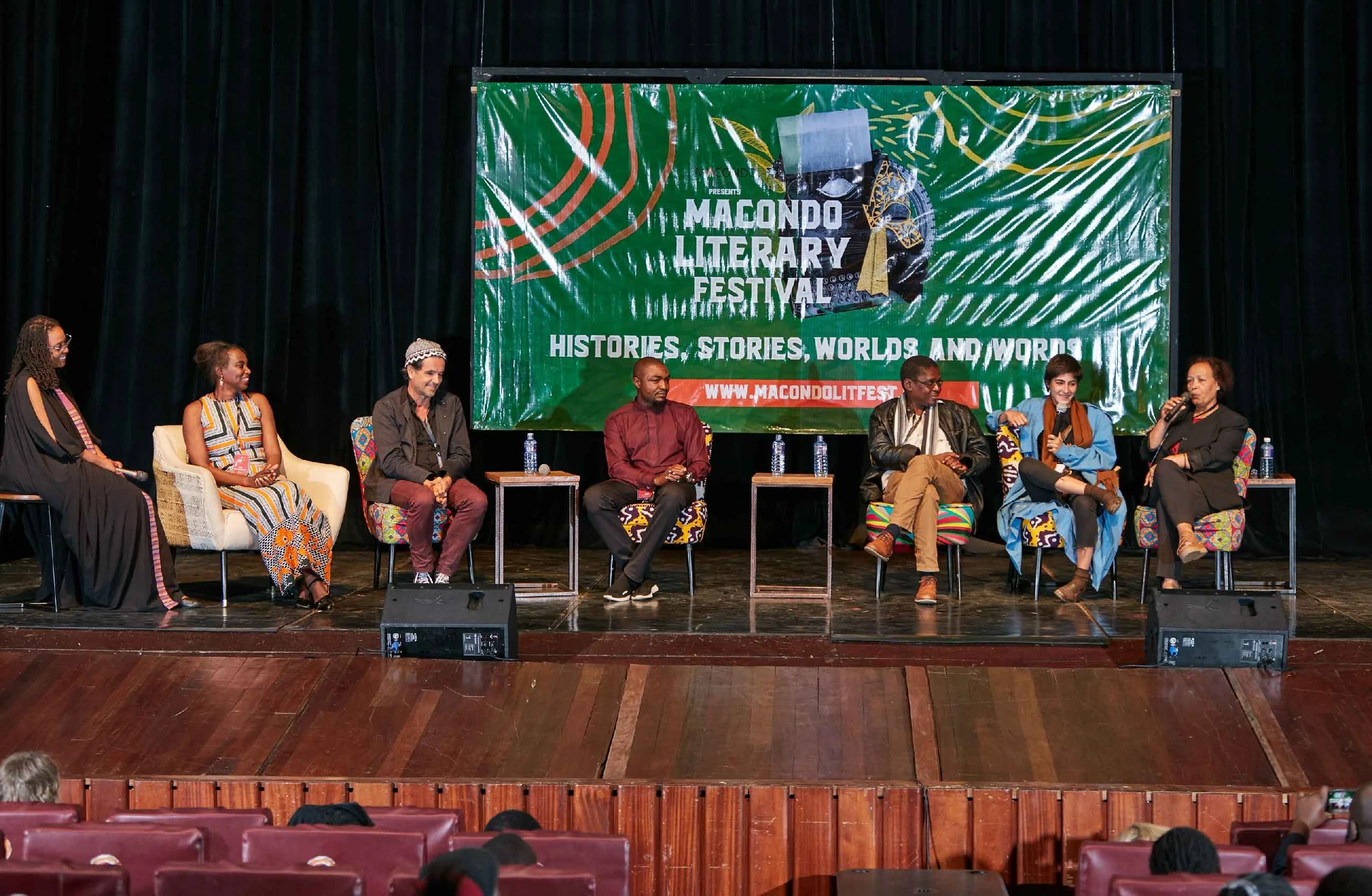 Panel discussion at the Macondo Literary Festival with seven diverse speakers seated on stage, engaging in conversation. The backdrop shows a large green banner with the festival name, theme 'Histories, Stories, Worlds, and Words,' and website.