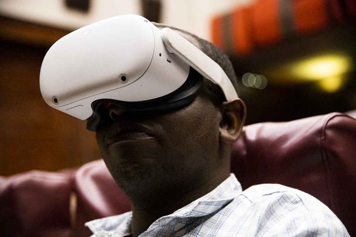 A man wearing a white virtual reality headset, sitting on a red chair in an indoor setting with blurred background lights.