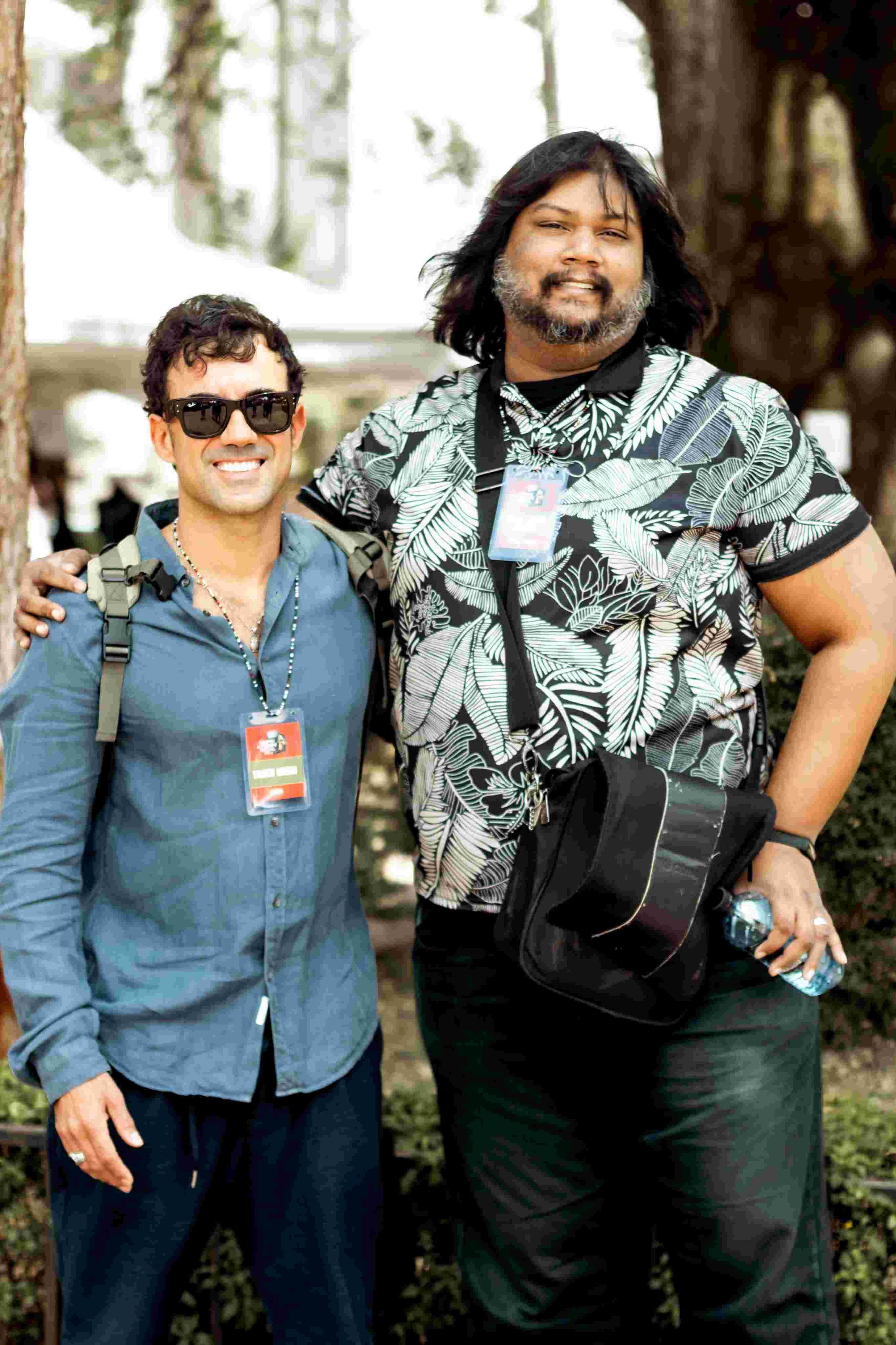 Two men standing outdoors, smiling, with trees in the background. The man on the left is wearing sunglasses, a blue button-up shirt, and has a backpack. The man on the right has long dark hair, a beard, and is wearing a patterned shirt, holding a wat
