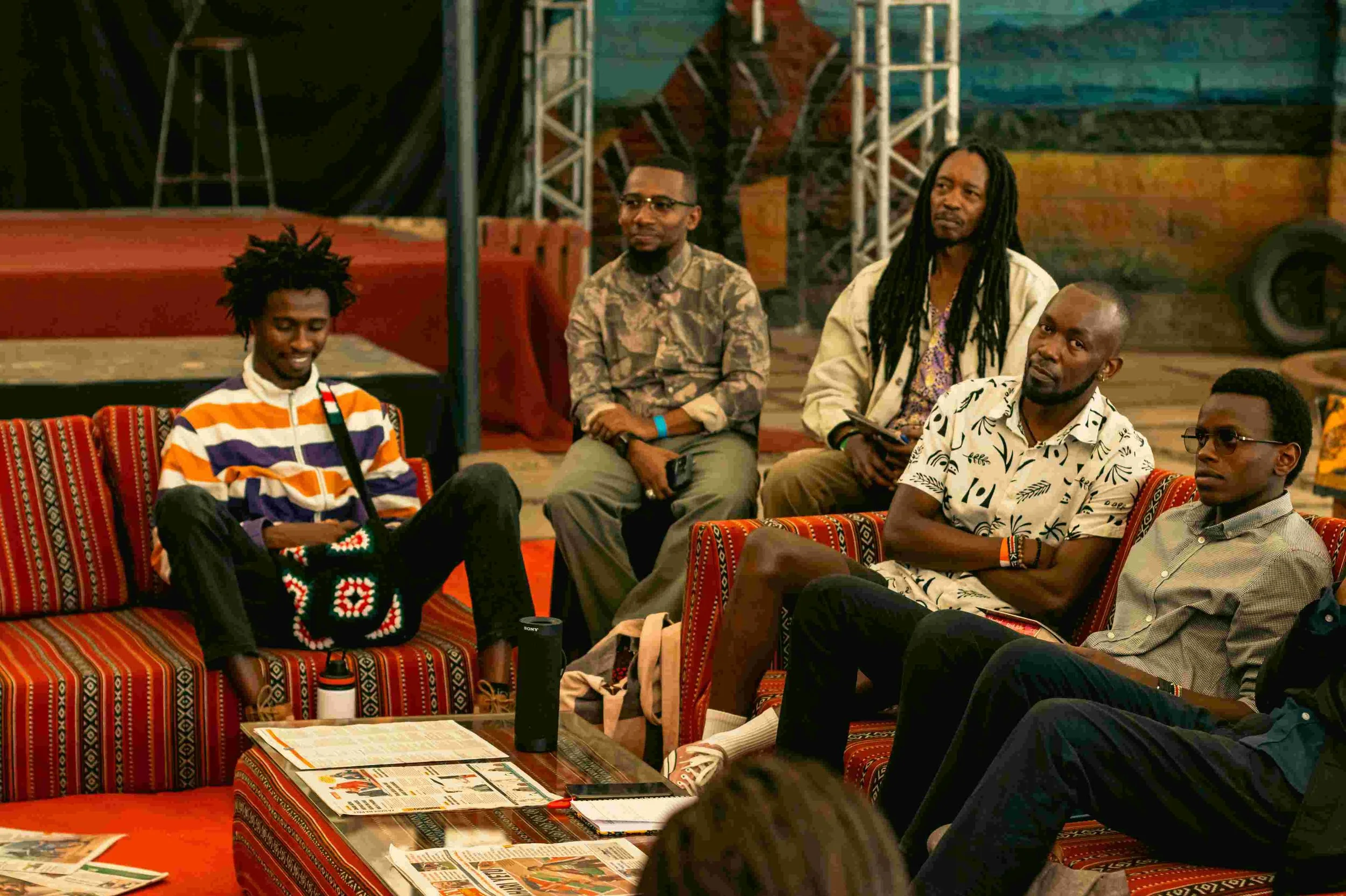 Group of six men sitting on colorful patterned couches in a discussion or meeting setting, surrounded by tables with newspapers and personal items, in a room with a stage and industrial decor.