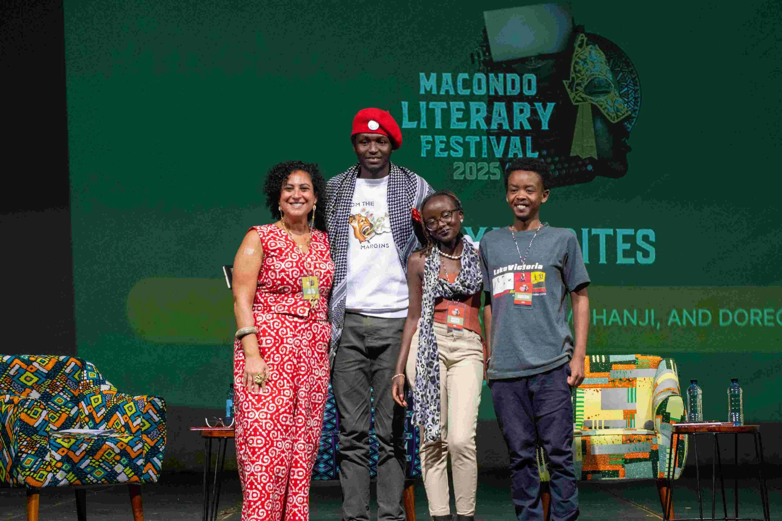 Four diverse people standing on stage at the Macondo Literary Festival 2025, with a large green screen behind them displaying text and logo, and two patterned armchairs with glasses of water on tables on either side of the stage.