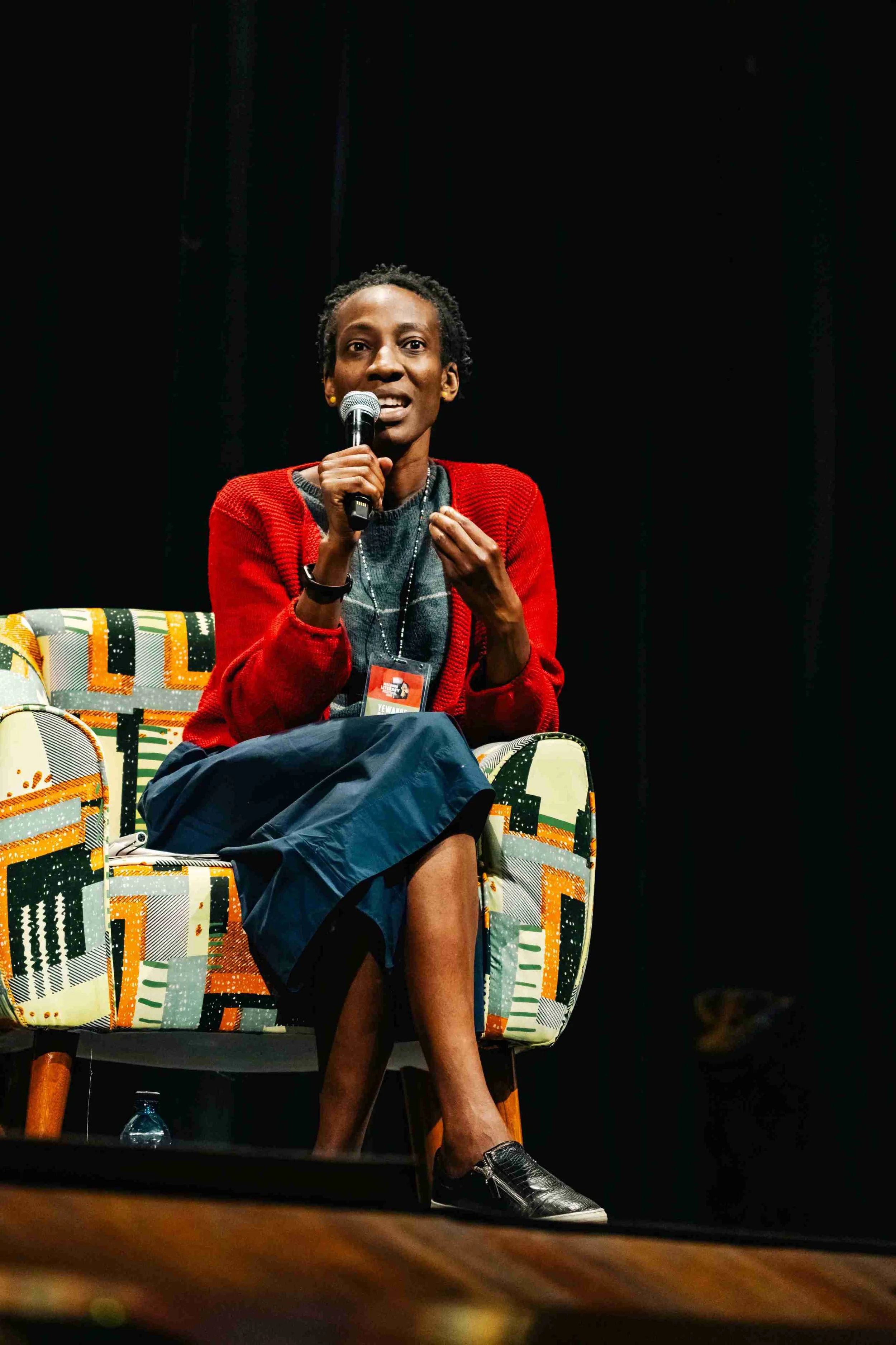 A woman speaking into a microphone while sitting on a patterned chair, wearing a red cardigan, black skirt, and black shoes, with a black background behind her.