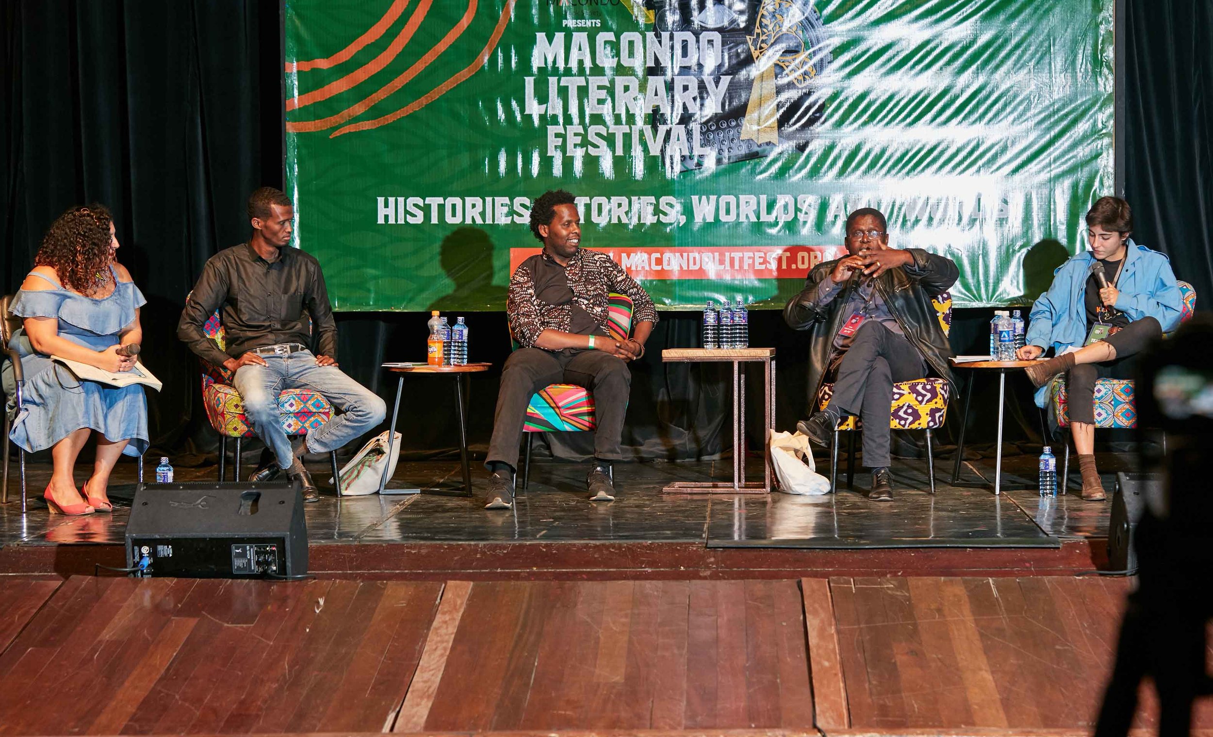 A panel discussion with five people seated on stage in front of a green banner at the Macondo Literary Festival. The panelists are engaged in conversation, with two women and three men, each with water bottles and microphones.