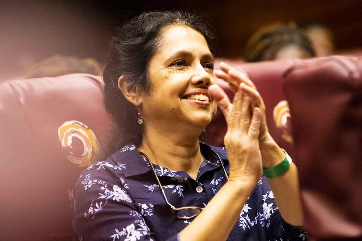 A woman claps and smiles in an indoor setting with maroon upholstered chairs.