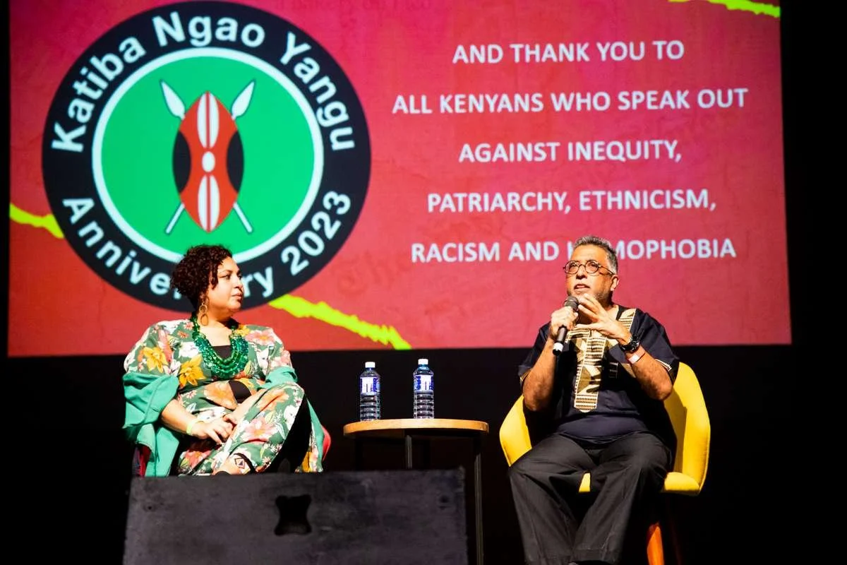 A woman and a man sitting on stage during a discussion at Katiba Nao Yangu's 2023 anniversary event, with a large screen behind them displaying a logo and a message of appreciation to Kenyans speaking out against inequity, patriarchy, ethnocism, raci