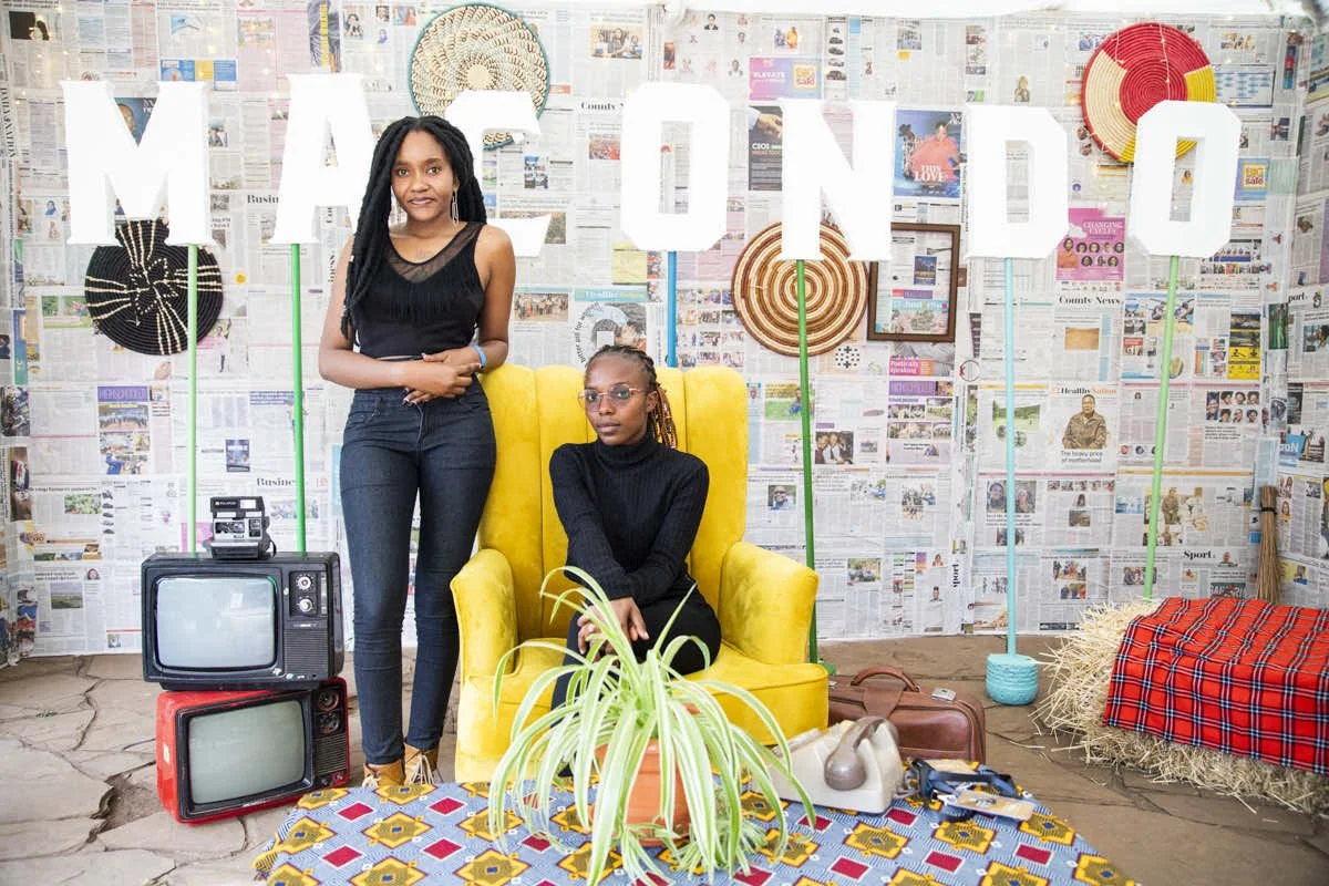 Two women in a colorful, eclectic room with a background of newspaper-covered walls arranged to spell 'AMONI' with large white letters held by green poles. One woman with long black dreadlocks stands next to a seated woman with glasses, both dressed 