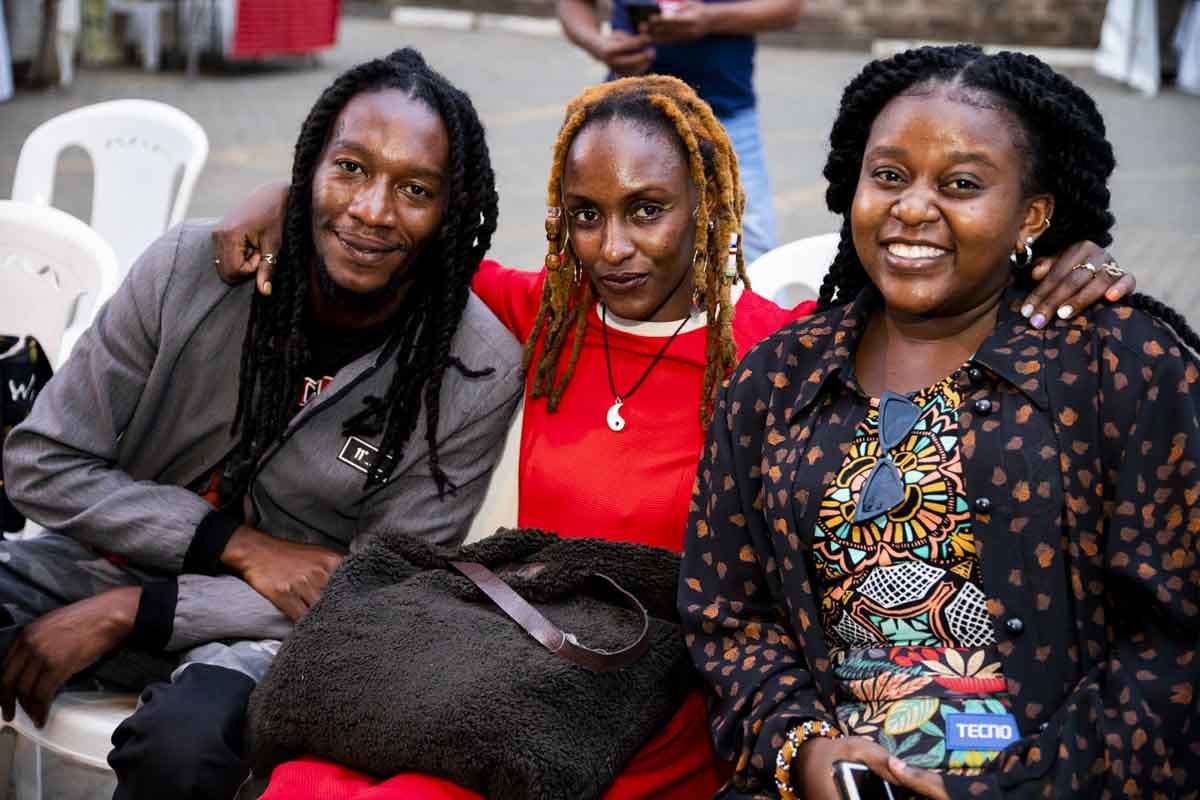 Three women sitting together outdoors, smiling, with white chairs and a person standing in the background.