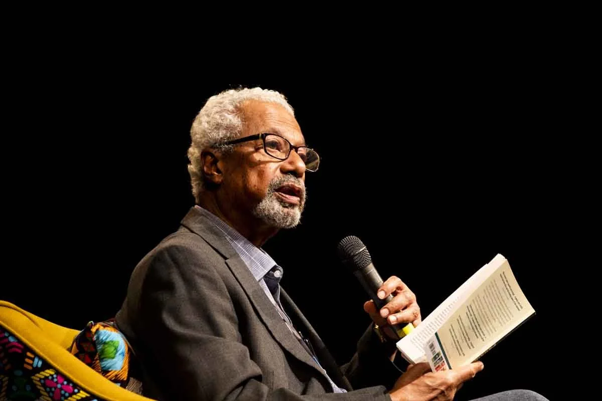 An elderly man with glasses, gray hair, and a beard, sitting on a yellow chair, wearing a blazer, holding a microphone, and reading a book on stage with a black background.