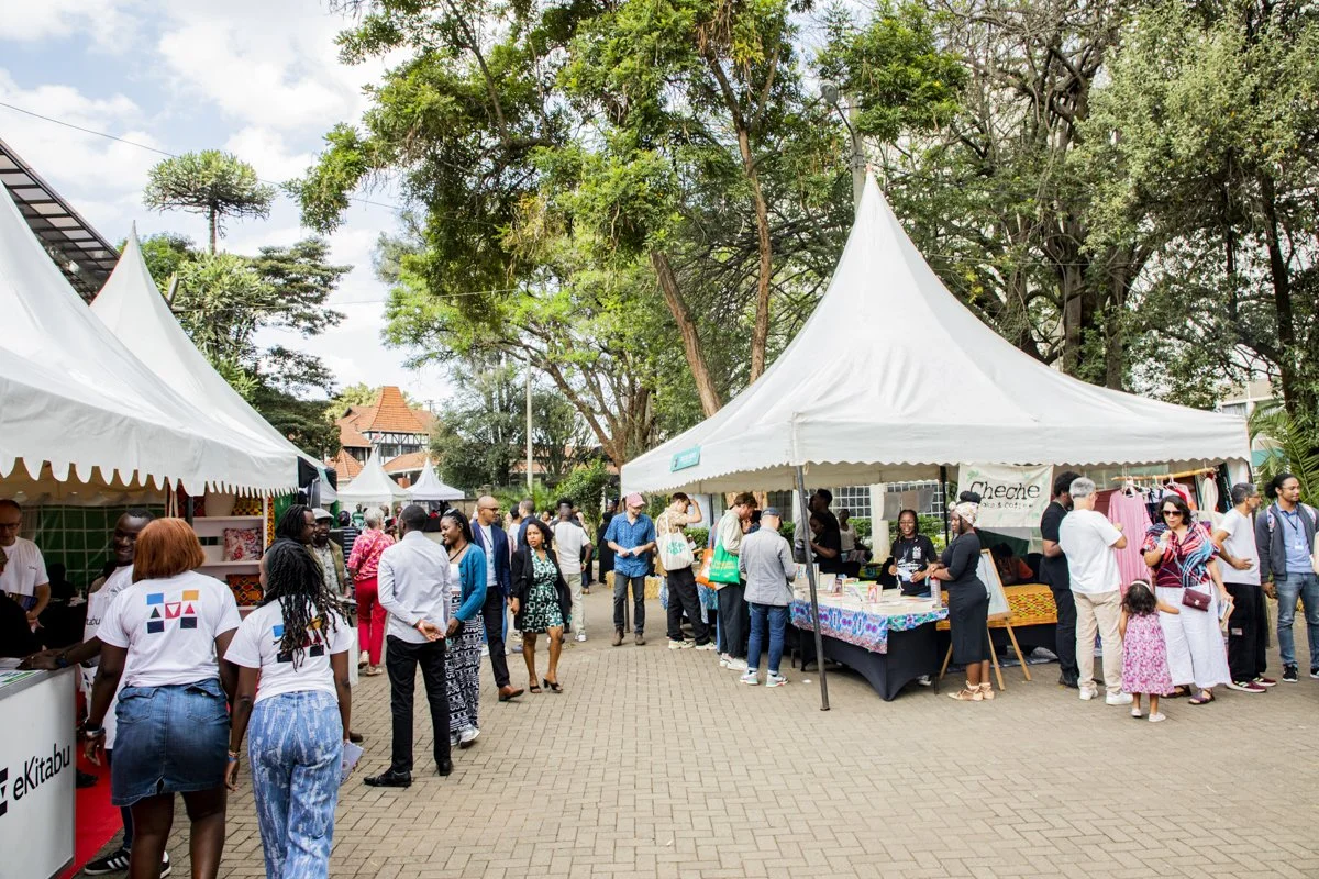 People browsing vendor booths at an outdoor market under trees with a cloudy sky in the background.