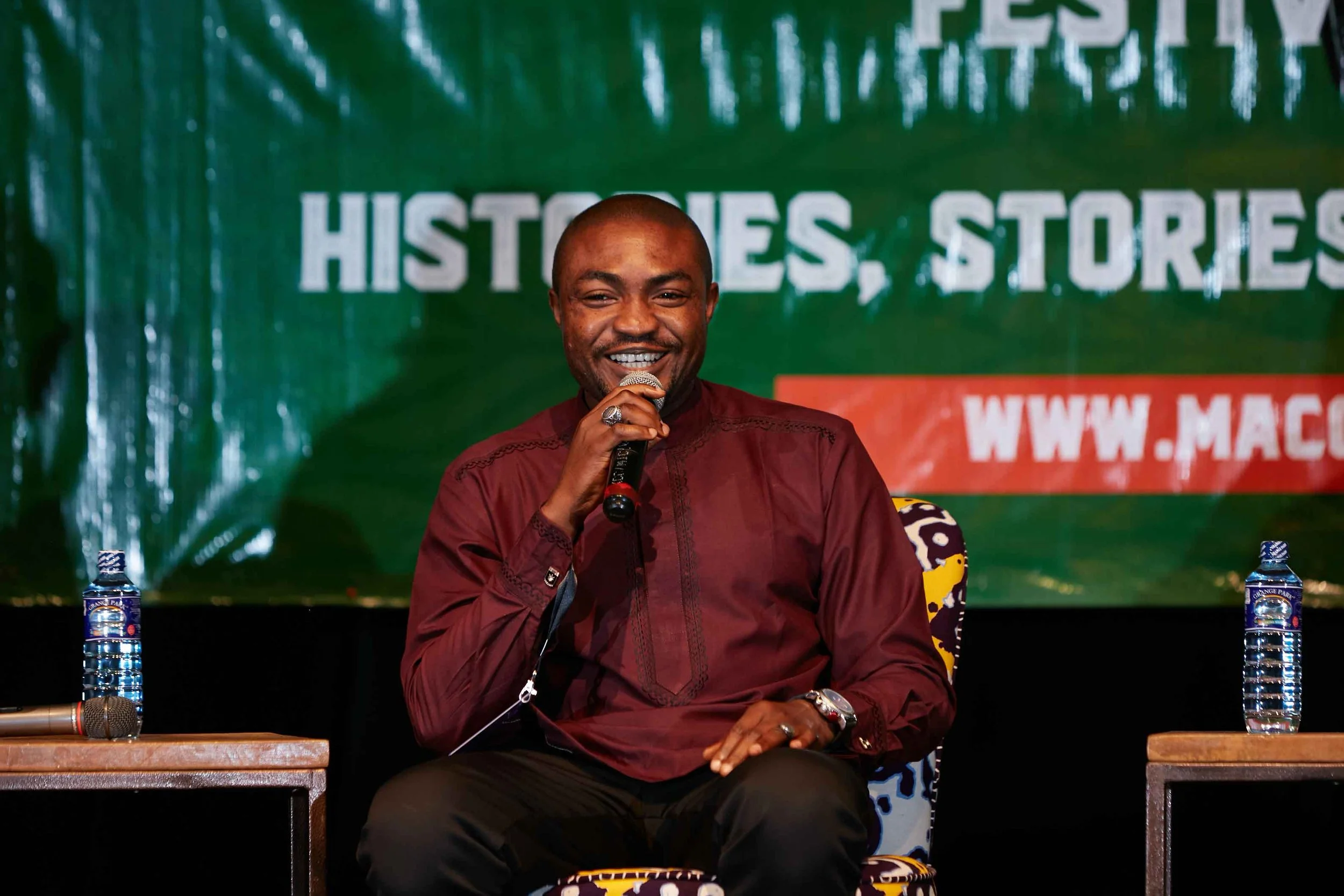 A smiling man sitting on a chair, holding a microphone, during a presentation or event. Behind him is a green banner with white text that reads 'HISTORIES, STORIES' and a red URL partially visible. Two water bottles are on tables on either side of hi